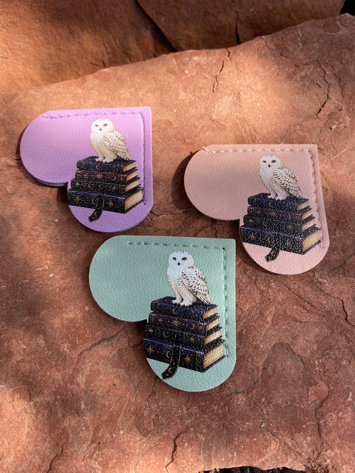 Three heart-shaped leather accessories with stitched edges, each featuring an owl sitting on a stack of books, displayed on a textured reddish-brown rock surface. The accessories come in lavender, pale pink, and mint green colorways.