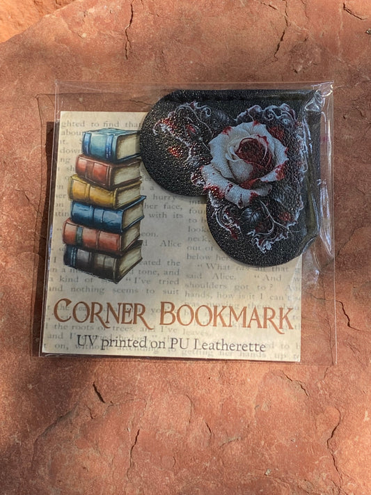 A packaged corner bookmark set on a textured terracotta surface. The set includes a rectangular card printed with vintage book stacks and the words 'CORNER BOOKMARK' and 'UV printed on PU Leatherette,' plus two oval leatherette corner pieces: one black with a white rose design and one black with a red rose design.