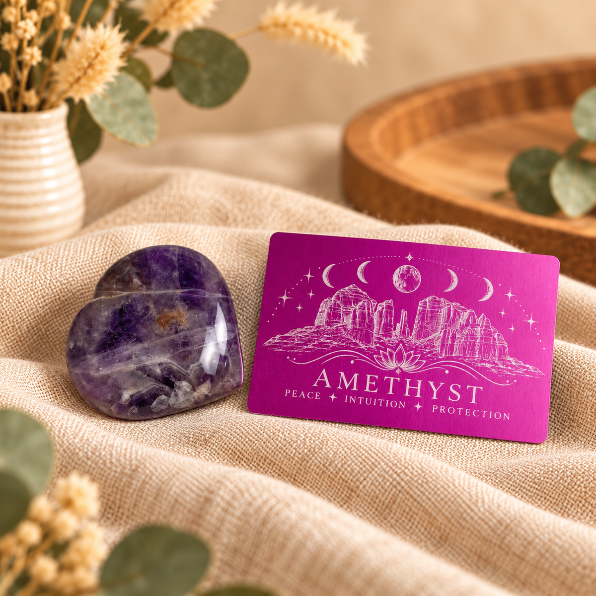 A purple amethyst crystal heart and a matching rectangular card labeled 'AMETHYST PEACE INTUITION PROTECTION' sit on beige fabric, with dried flowers and a wooden tray blurred in the background.