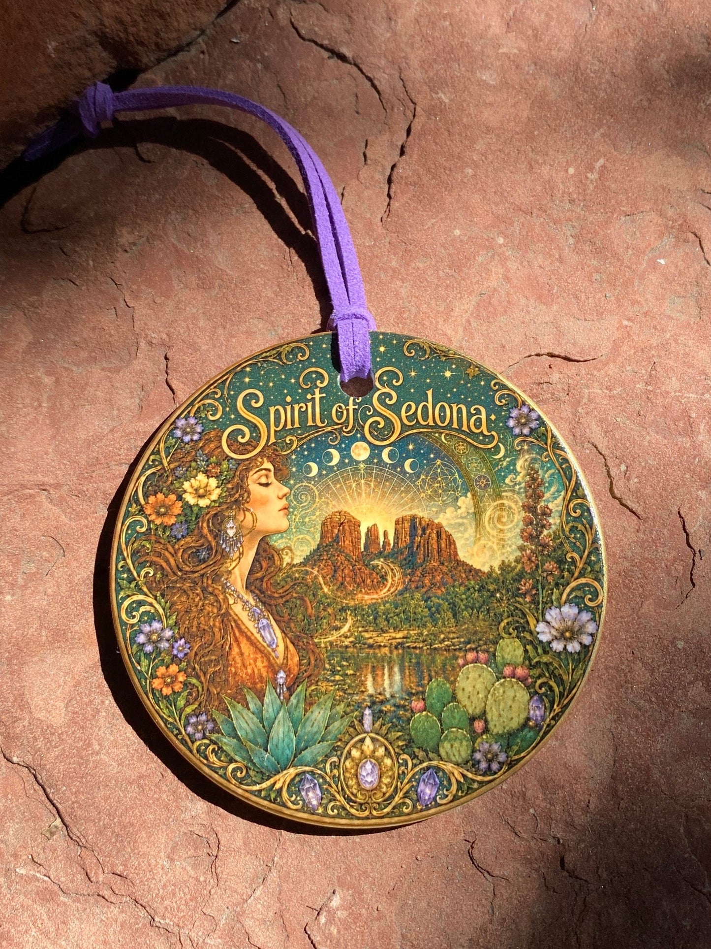 A round decorative ornament featuring an Art Nouveau style illustration of a woman in a forest with the text 'Spirit of Edona' and a purple hanging cord, placed on a textured reddish-brown stone surface.