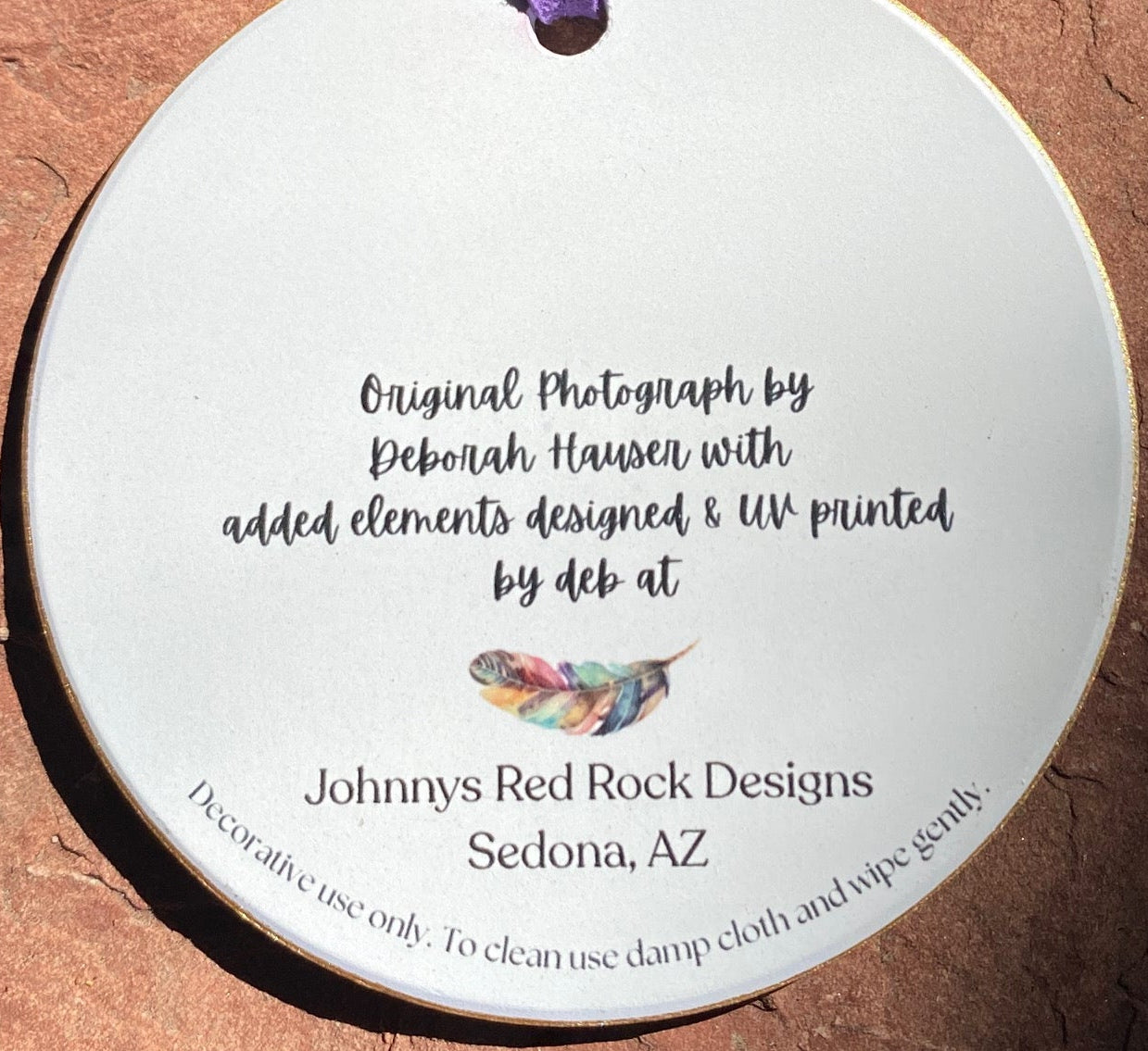 Back of ornament A round white ceramic tag or ornament with a purple hanging cord, photographed on a textured reddish-brown stone surface. The tag has black printed text and a small feather illustration.