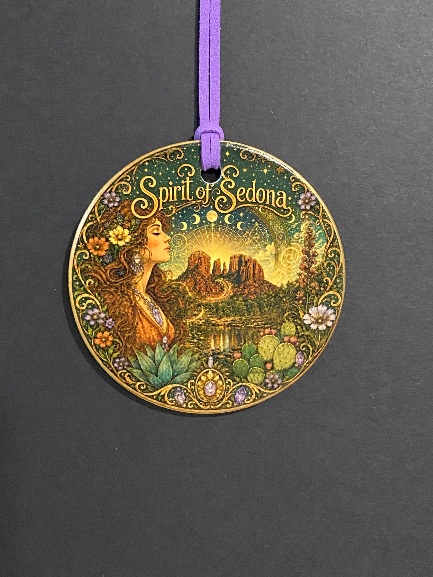 A round decorative medallion featuring an Art Nouveau style illustration of a woman in a forest with the text 'Spirit of Selena' on a dark gray background, attached to a purple cord.