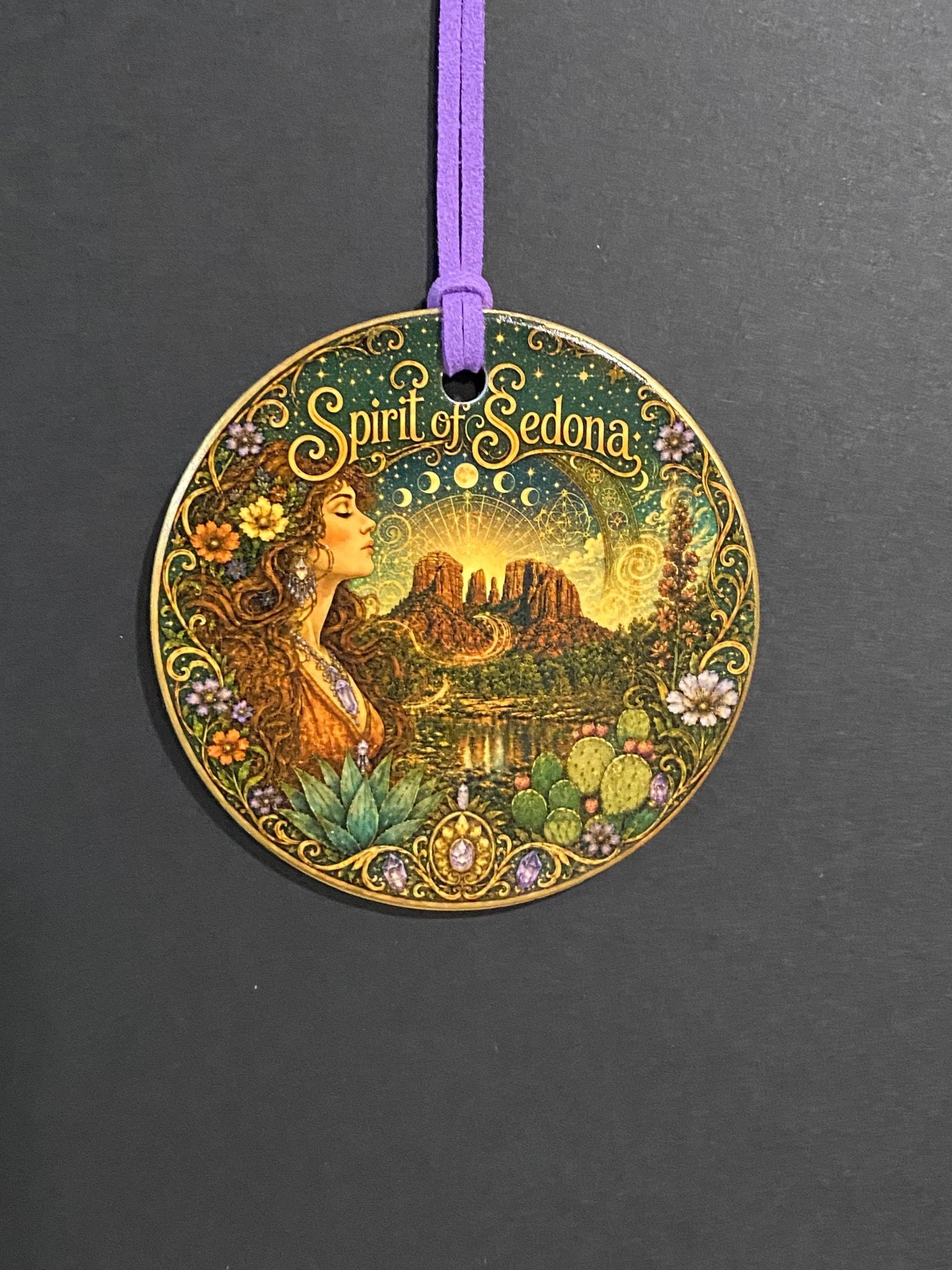 A round decorative medallion featuring an Art Nouveau style illustration of a woman in a forest with the text 'Spirit of Selena' on a dark gray background, attached to a purple cord.
