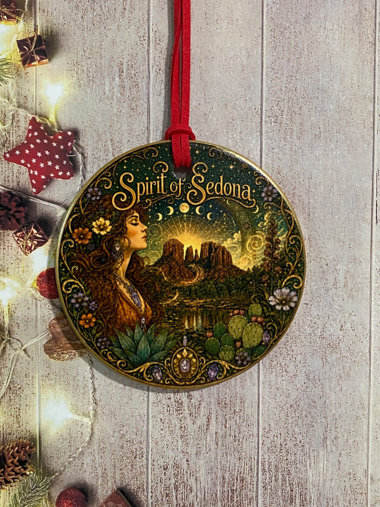 A round decorative ornament featuring an ornate fantasy illustration with the text 'Spirit of Sedona' and a red hanging cord, placed on a light wood surface with warm string lights and small holiday props around it.