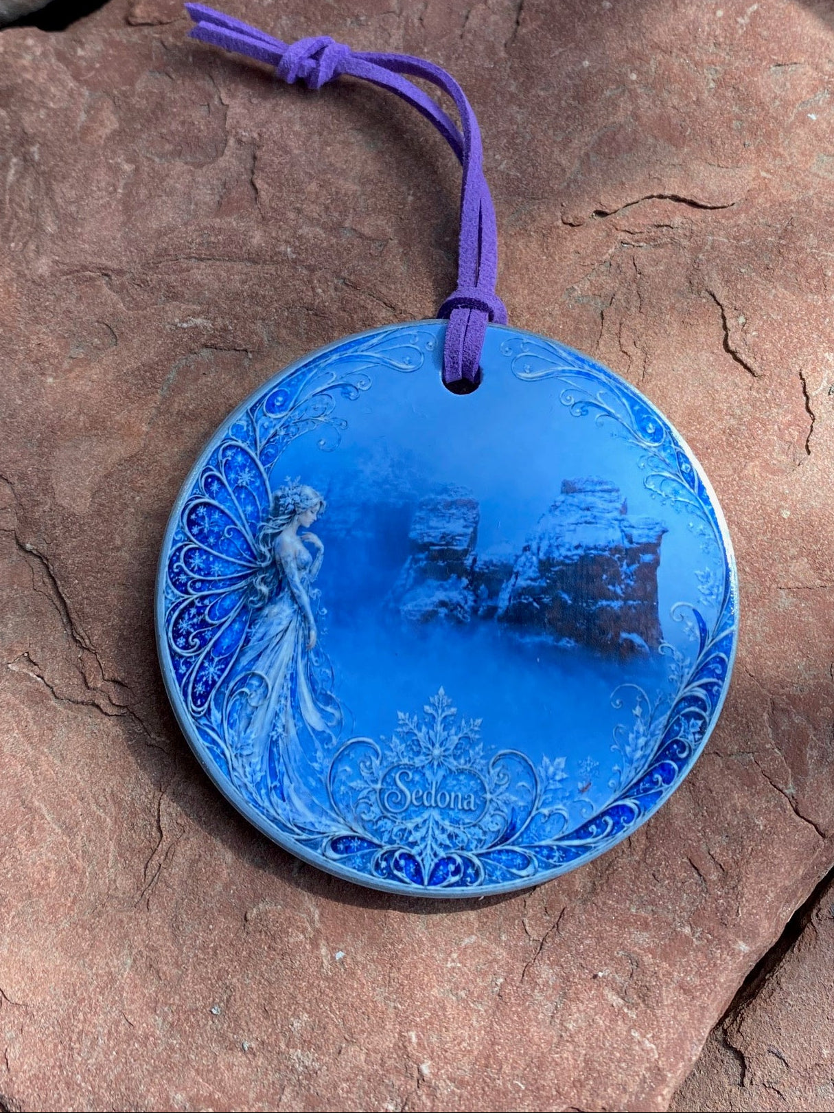 A round blue ceramic ornament with a purple hanging cord, featuring a printed fairy and cottage design, resting on a textured brown rock surface.