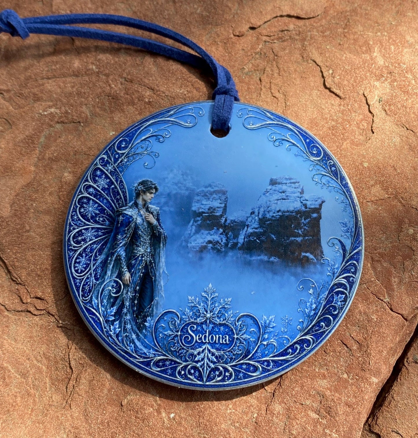 A round blue decorative plate with an engraved winter scene and a blue hanging cord, displayed on a textured brown stone surface.