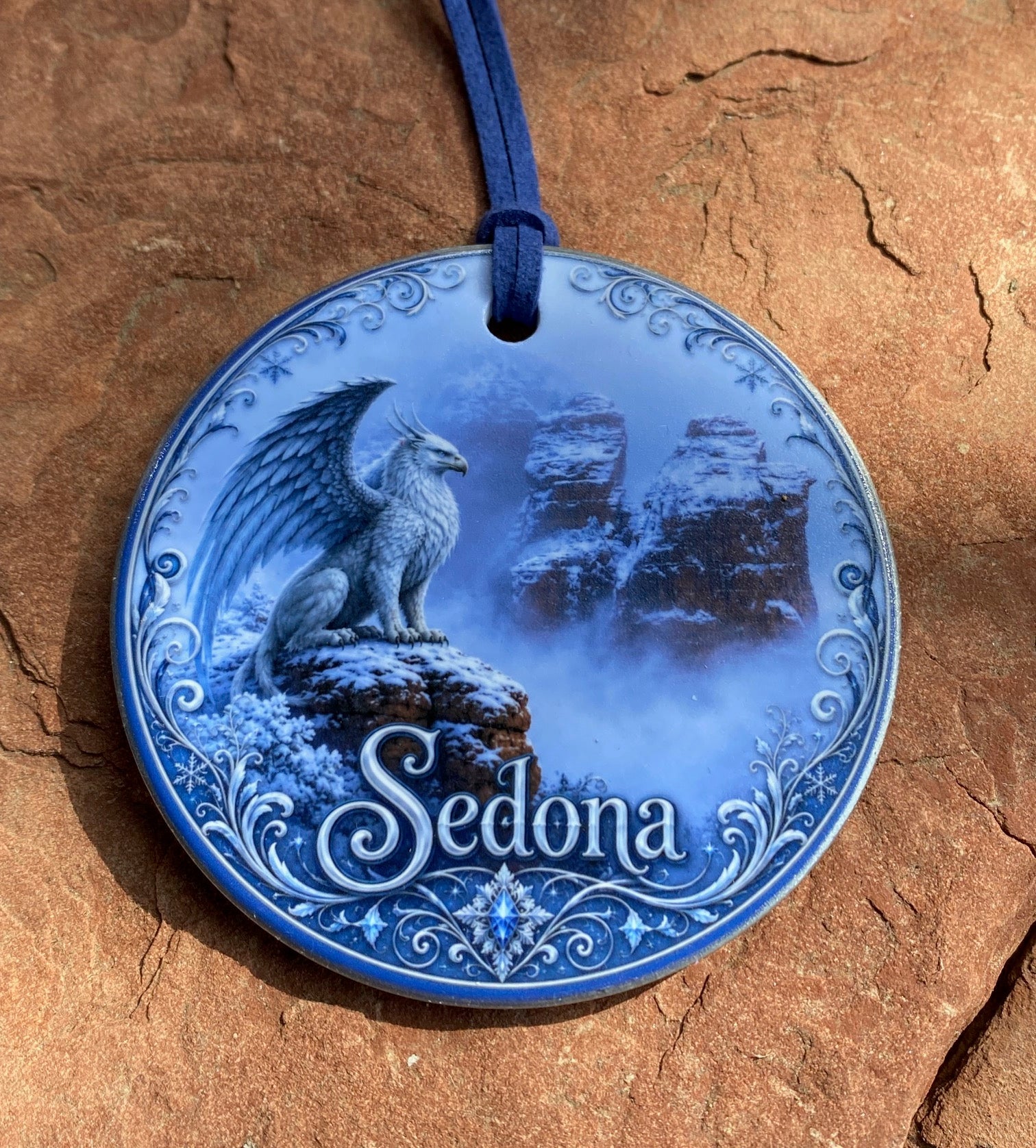 A round blue decorative plaque or ornament featuring an illustrated winged figure, ornate scrollwork, and the word 'Sedona' on a textured brown rock surface.