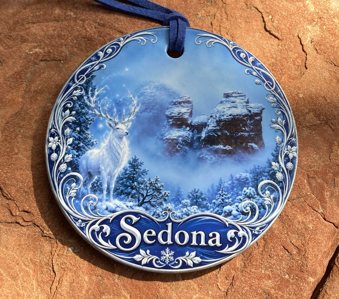 A round ceramic plate ornament featuring a blue and white winter landscape with the name 'Sedonas' on a blue side panel, hanging against a textured brown rock surface.