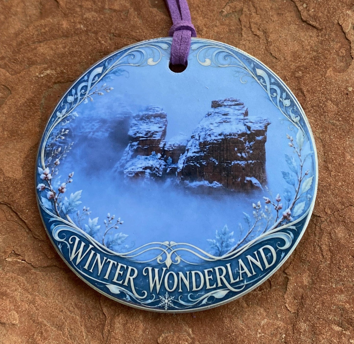 A round ceramic Christmas ornament featuring a blue winter forest scene with the text 'Winter Wonderland' on the rim, complete with a purple hanging cord, displayed on a textured brown stone surface.