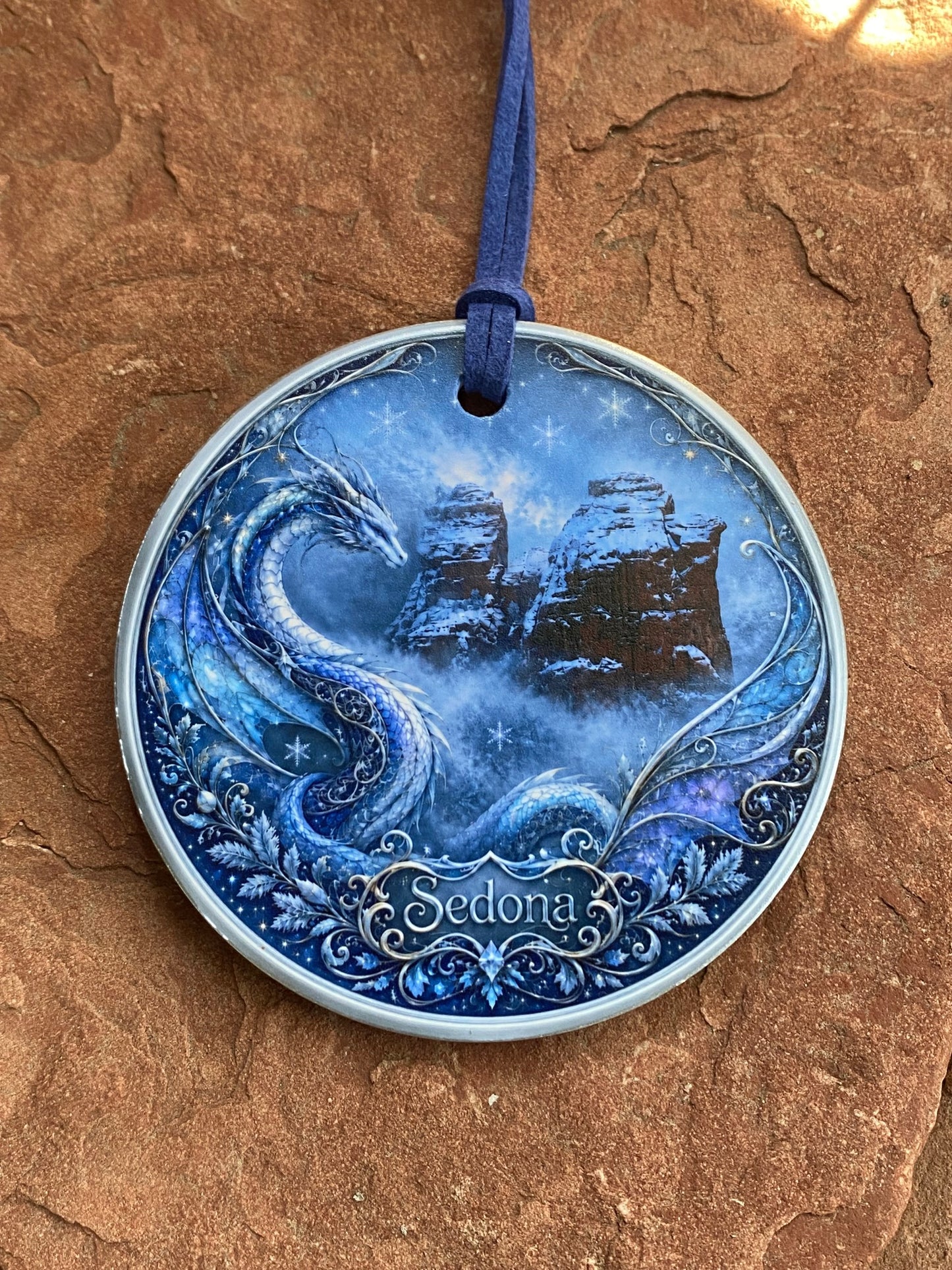 A round decorative medallion or pendant with a blue fantasy illustration of a dragon and castle, resting on a textured brown rock surface. A blue cord is threaded through a hole on the left side.