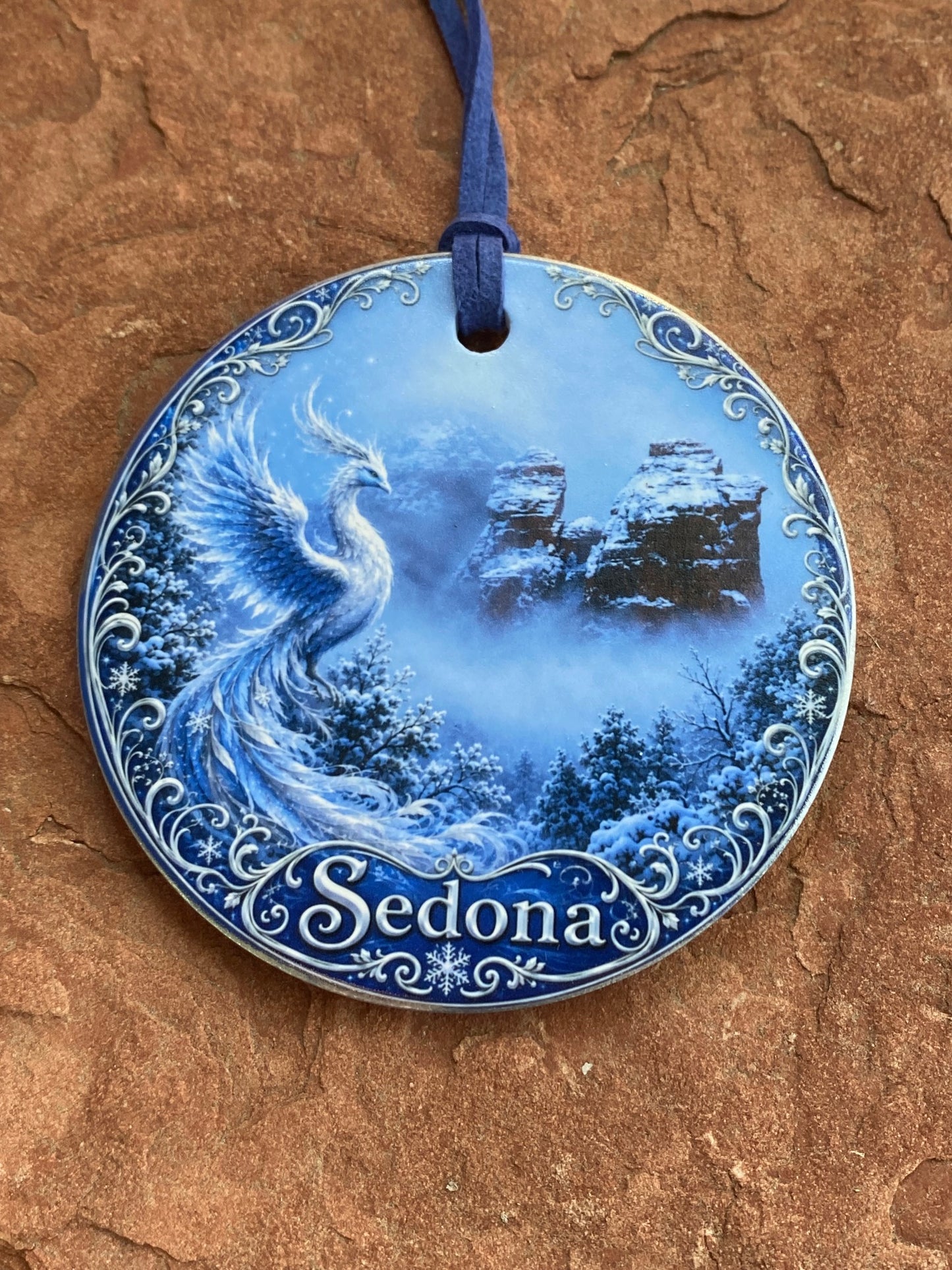 A round decorative plate or ornament with a blue and white seascape illustration and the name 'Sedona' on the right side, photographed against a textured brown stone surface. A dark blue ribbon loop is attached on the left.