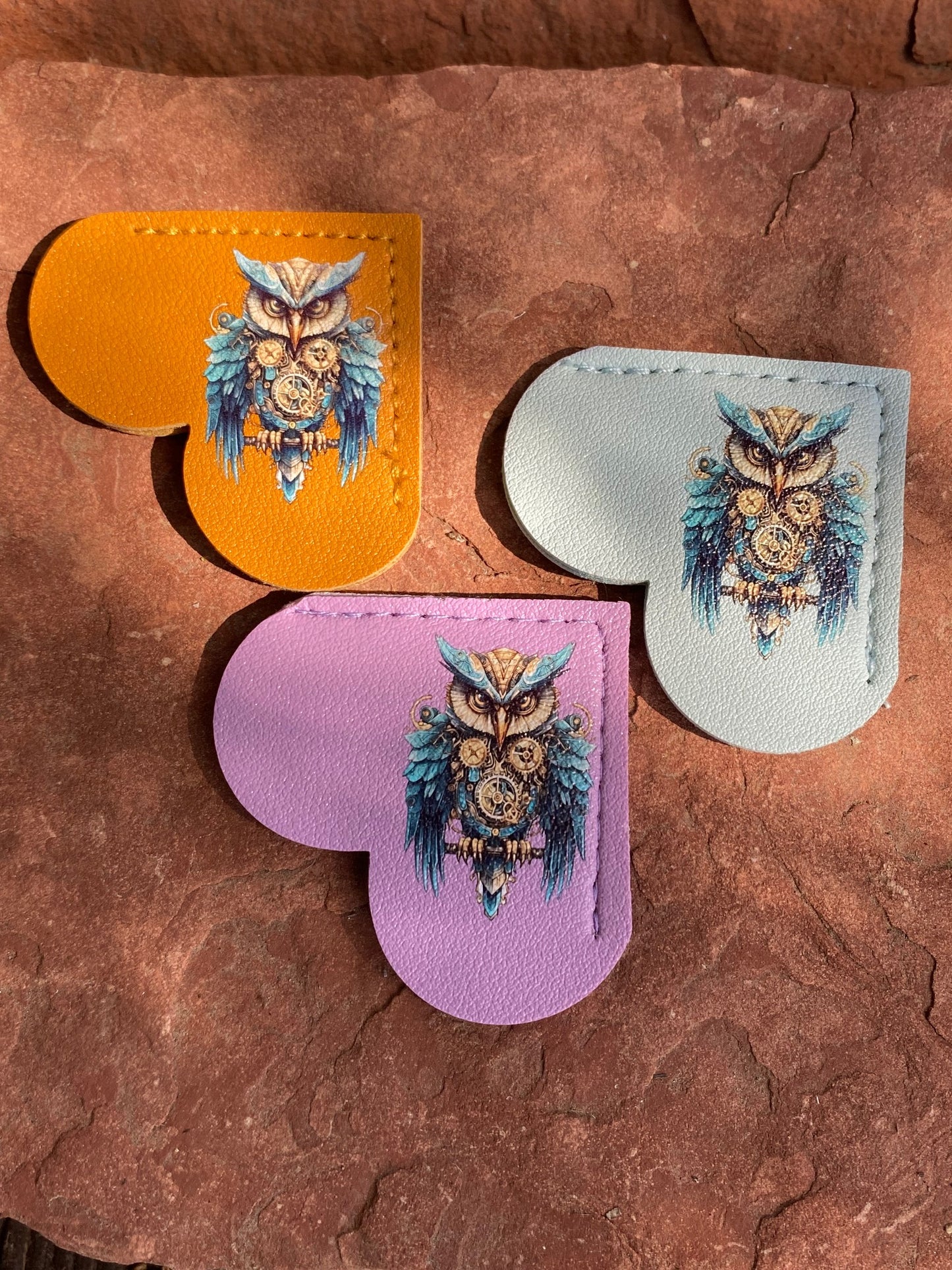 Three heart-shaped leather accessories with a detailed owl illustration, shown in orange, light blue, and lavender on a textured brown rock surface.