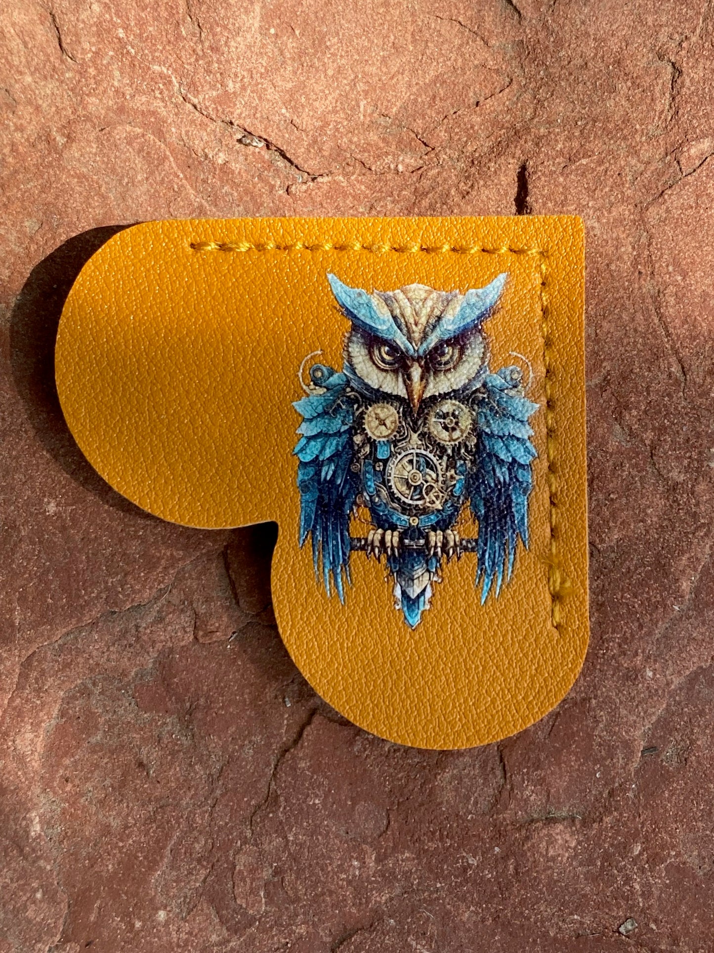 A mustard yellow leather pouch with a cutout, featuring a detailed illustration of a blue and silver owl on the front, displayed on a textured brown stone surface.