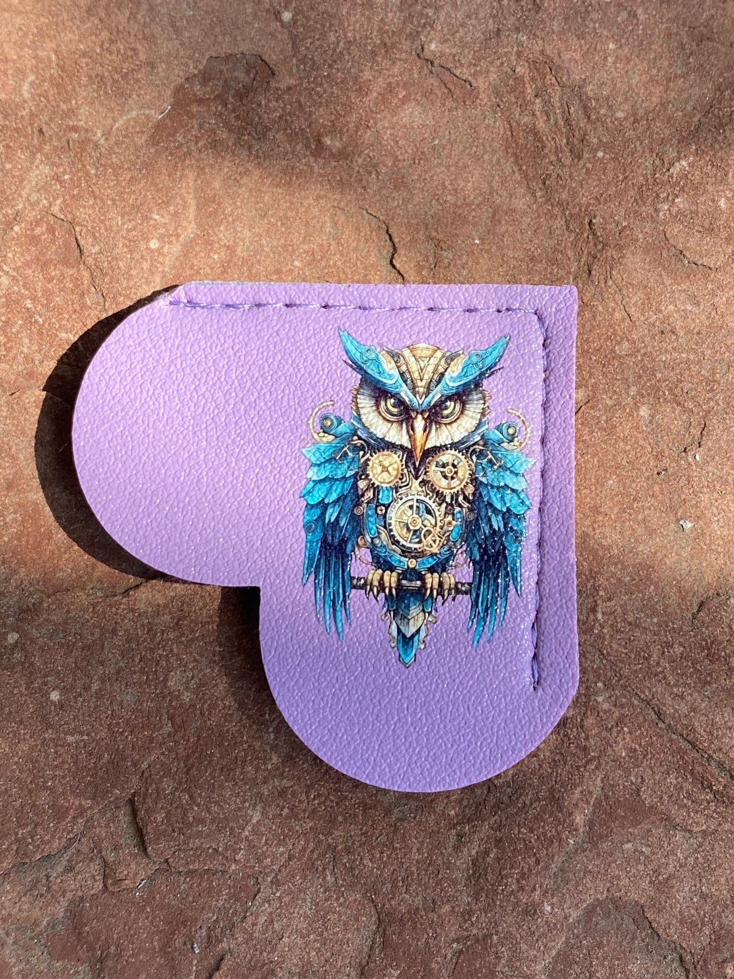 A lavender purple leather heart-shaped accessory with a stitched border and a detailed steampunk owl illustration in blue, gold, and white, displayed on a textured brown stone surface.
