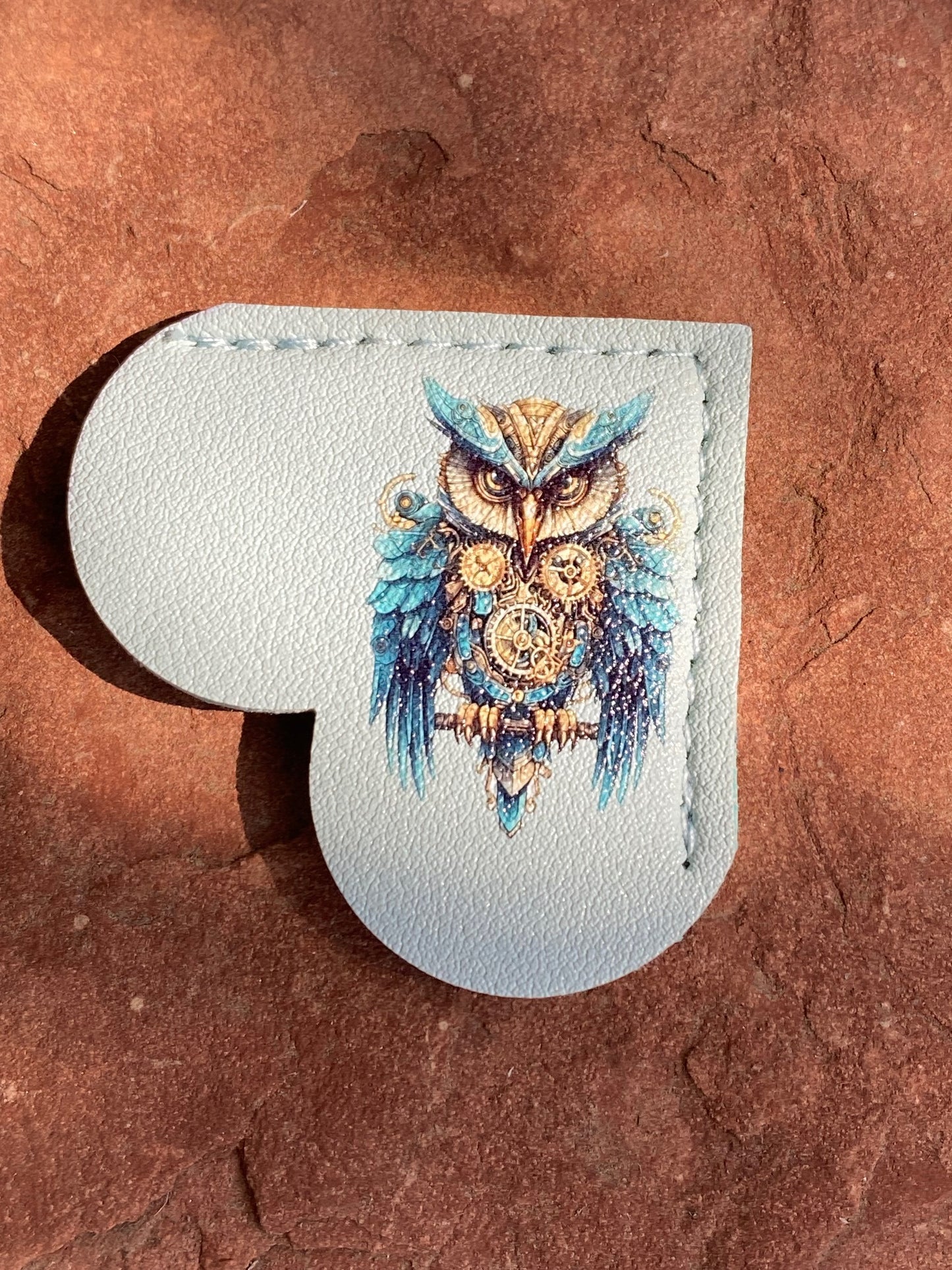 A heart-shaped light blue leather accessory featuring a detailed illustration of a blue and gold owl, displayed on a textured reddish-brown stone surface.