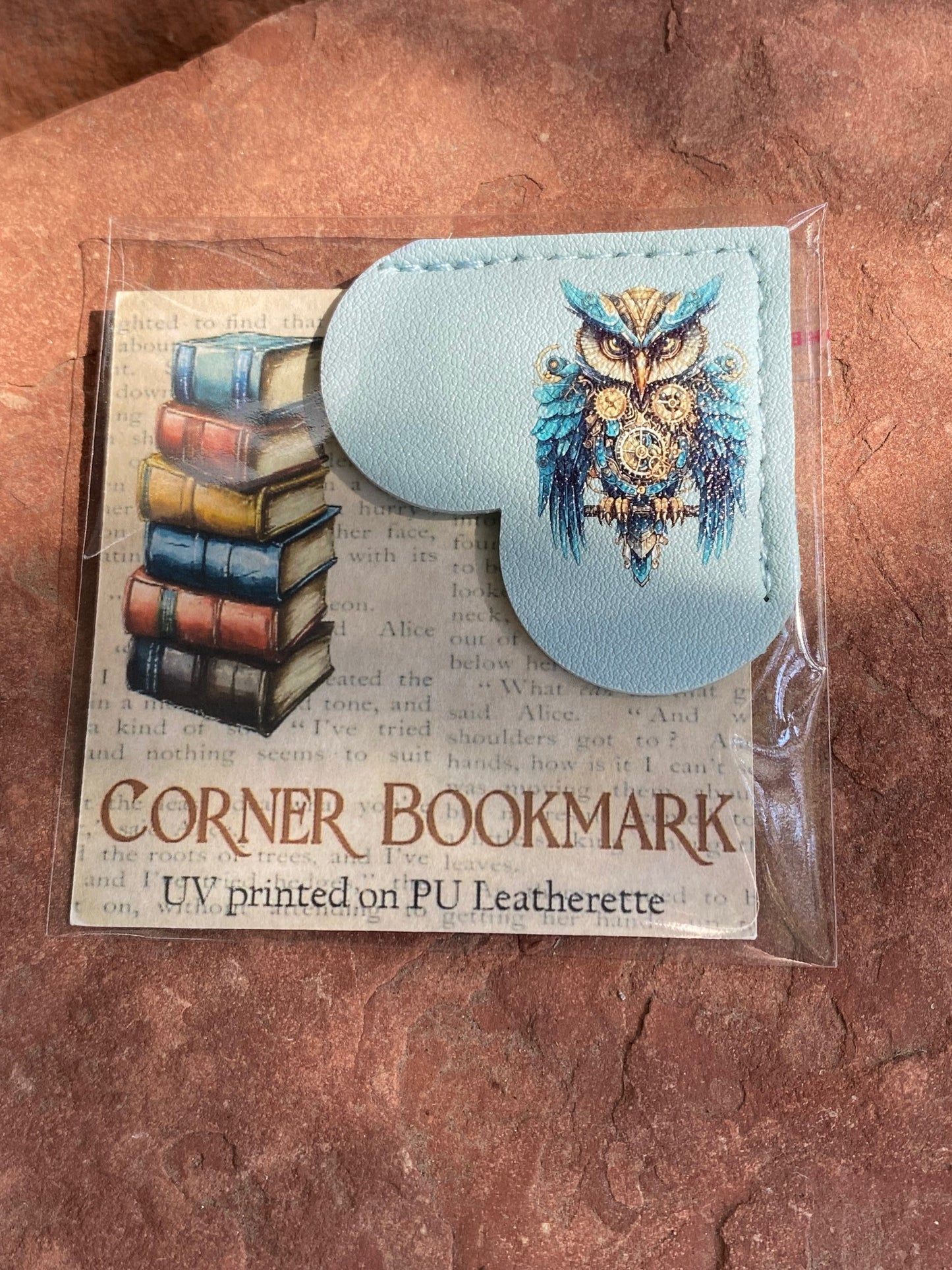A packaged corner bookmark on a textured brown surface. The bookmark is made of light blue PU leatherette and features a stitched heart shape with a printed owl design, alongside a printed insert card showing books and the text 'CORNER BOOKMARK' and 'UV printed on PU Leatherette'.