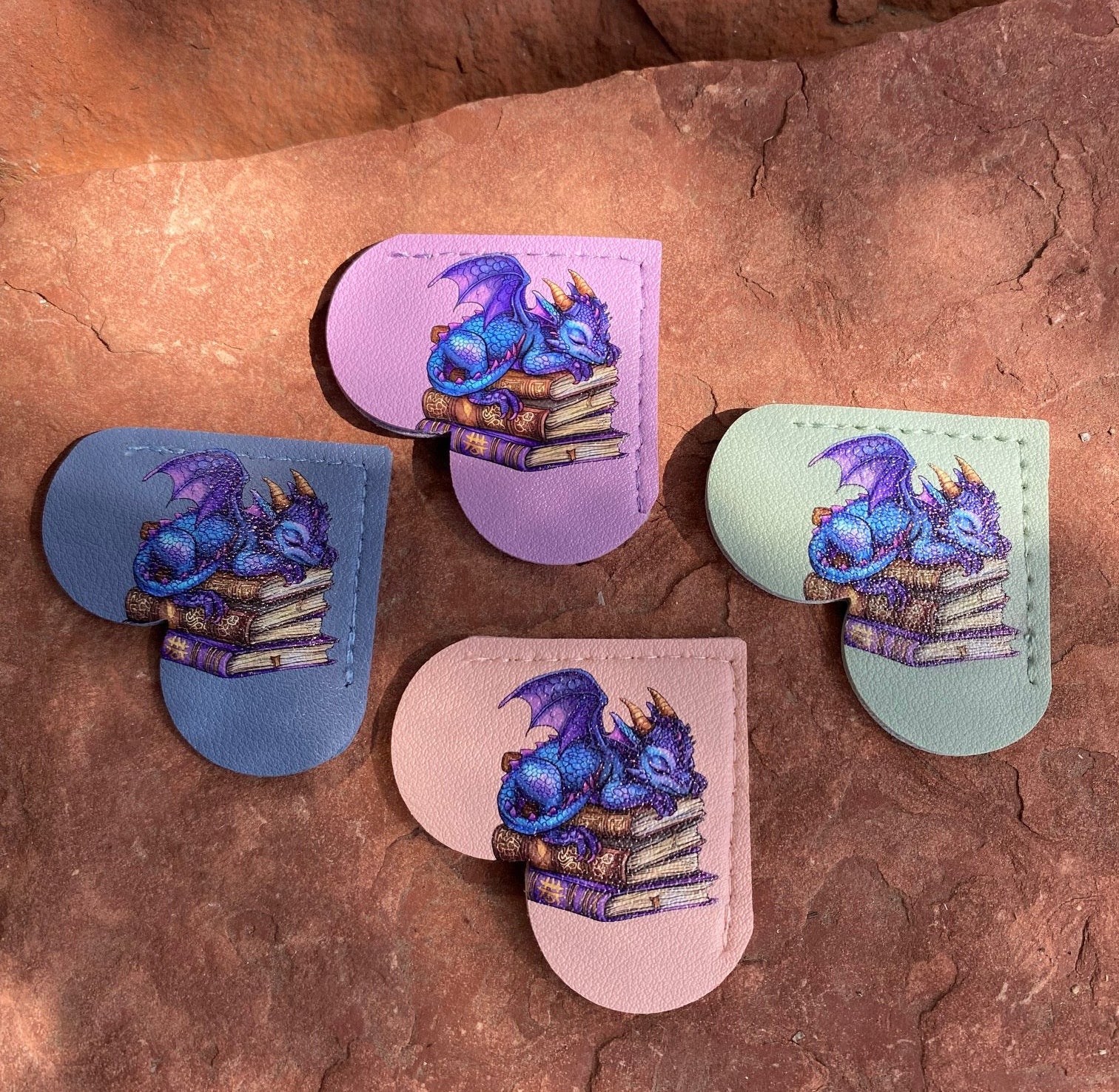Four heart-shaped leather keychains featuring a blue dragon sleeping on a stack of books, shown in pastel purple, blue, pink, and mint green on a textured reddish-brown rock background.