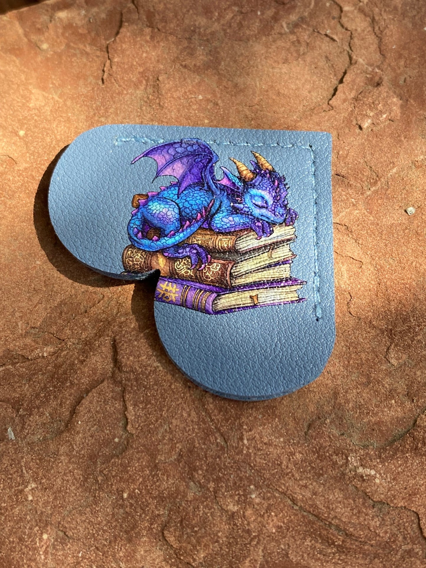 A light blue leather pouch with a heart-shaped cutout, featuring an illustration of a blue dragon sleeping on a stack of books. The pouch is placed on a textured brown stone surface.
