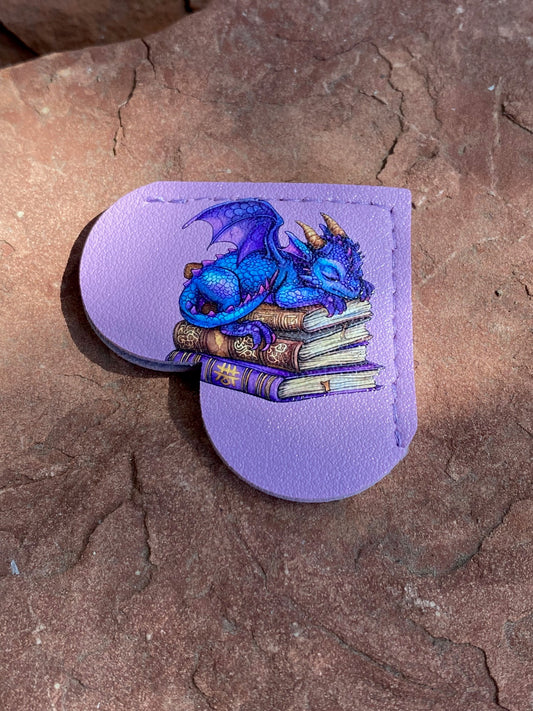 A lavender leather card holder or clutch featuring a detailed illustration of a blue dragon curled up on a stack of books, set against a textured brown stone background.