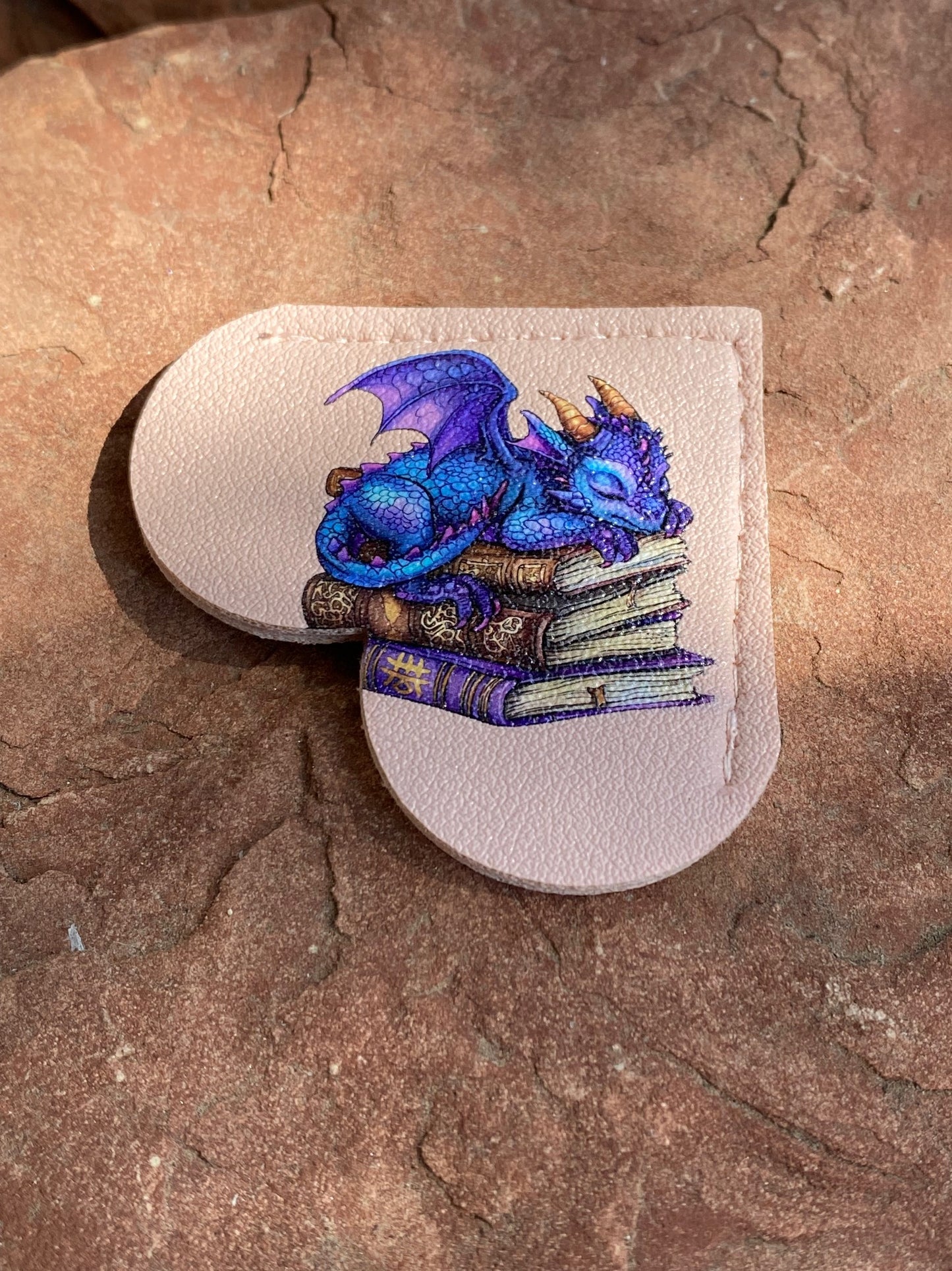 A custom embroidered patch featuring a blue and purple dragon sleeping on a stack of books, mounted on a light beige leather backing, placed on a textured brown stone surface.