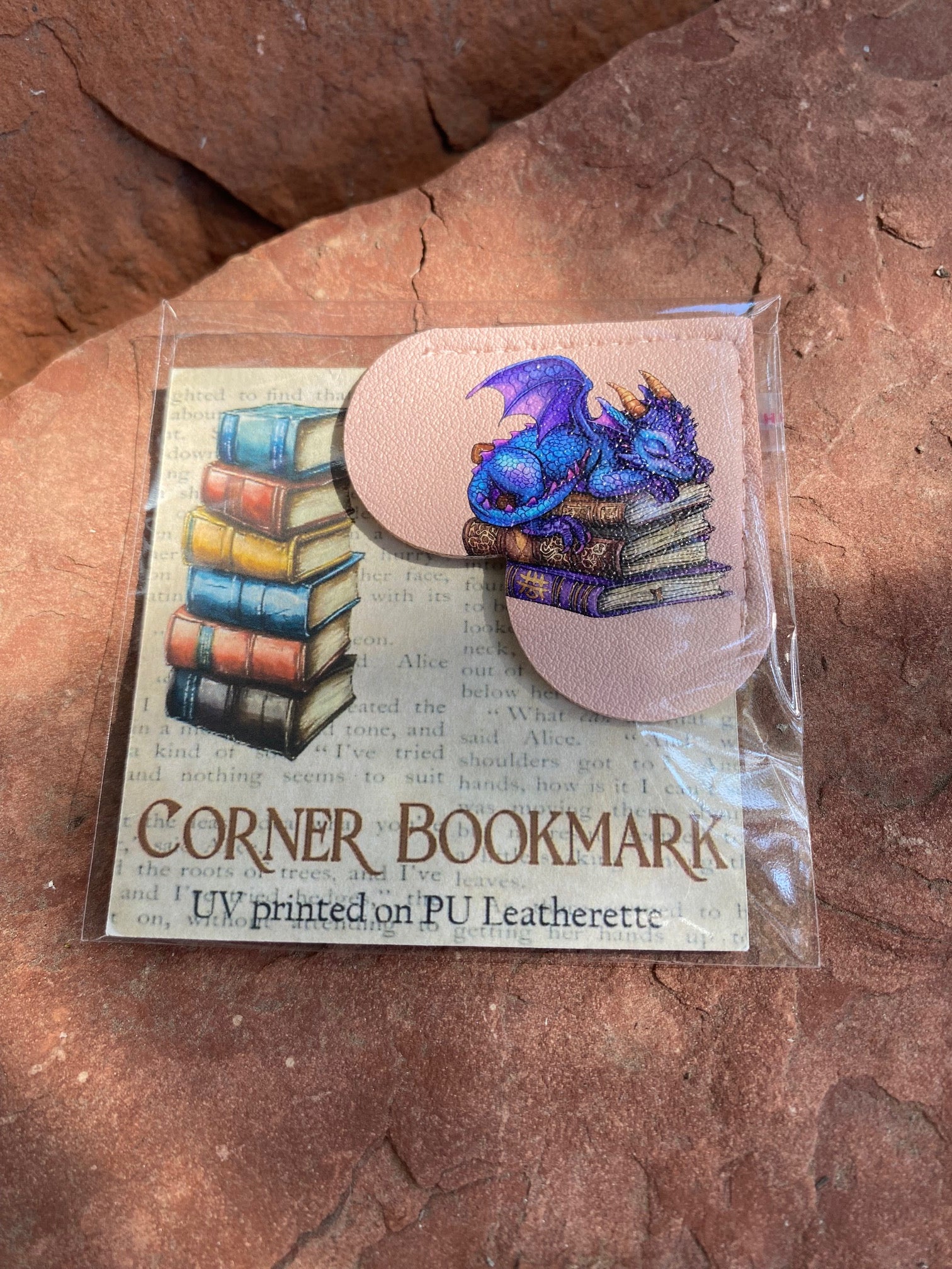 A packaged corner bookmark featuring a UV-printed illustration of a blue dragon curled over a stack of books, displayed on a textured reddish-brown rock surface.