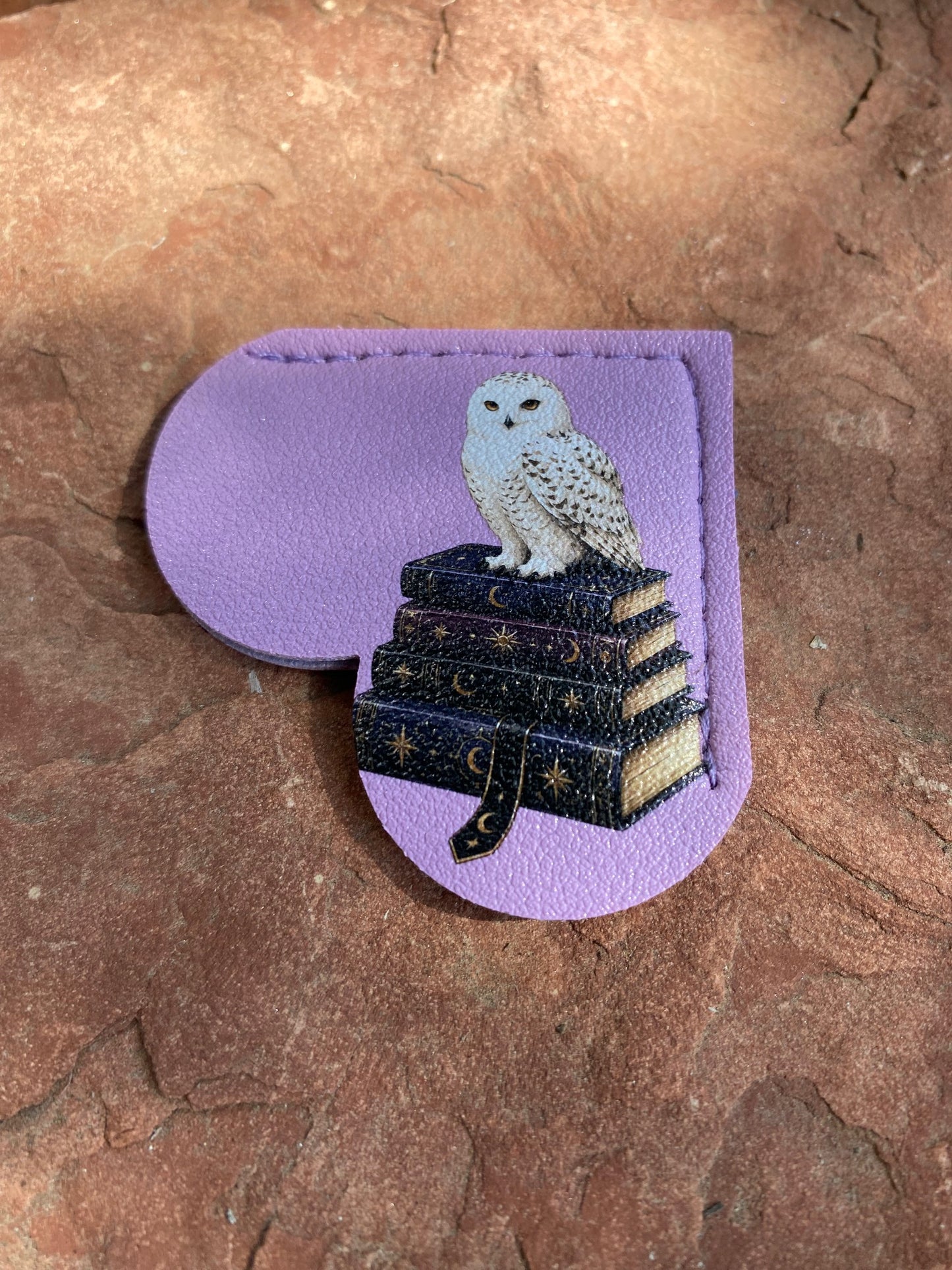 A lavender leather pouch with a die-cut shape, featuring an illustration of a white owl perched on a stack of dark, star-spangled books. The pouch is displayed on a textured brown stone surface.