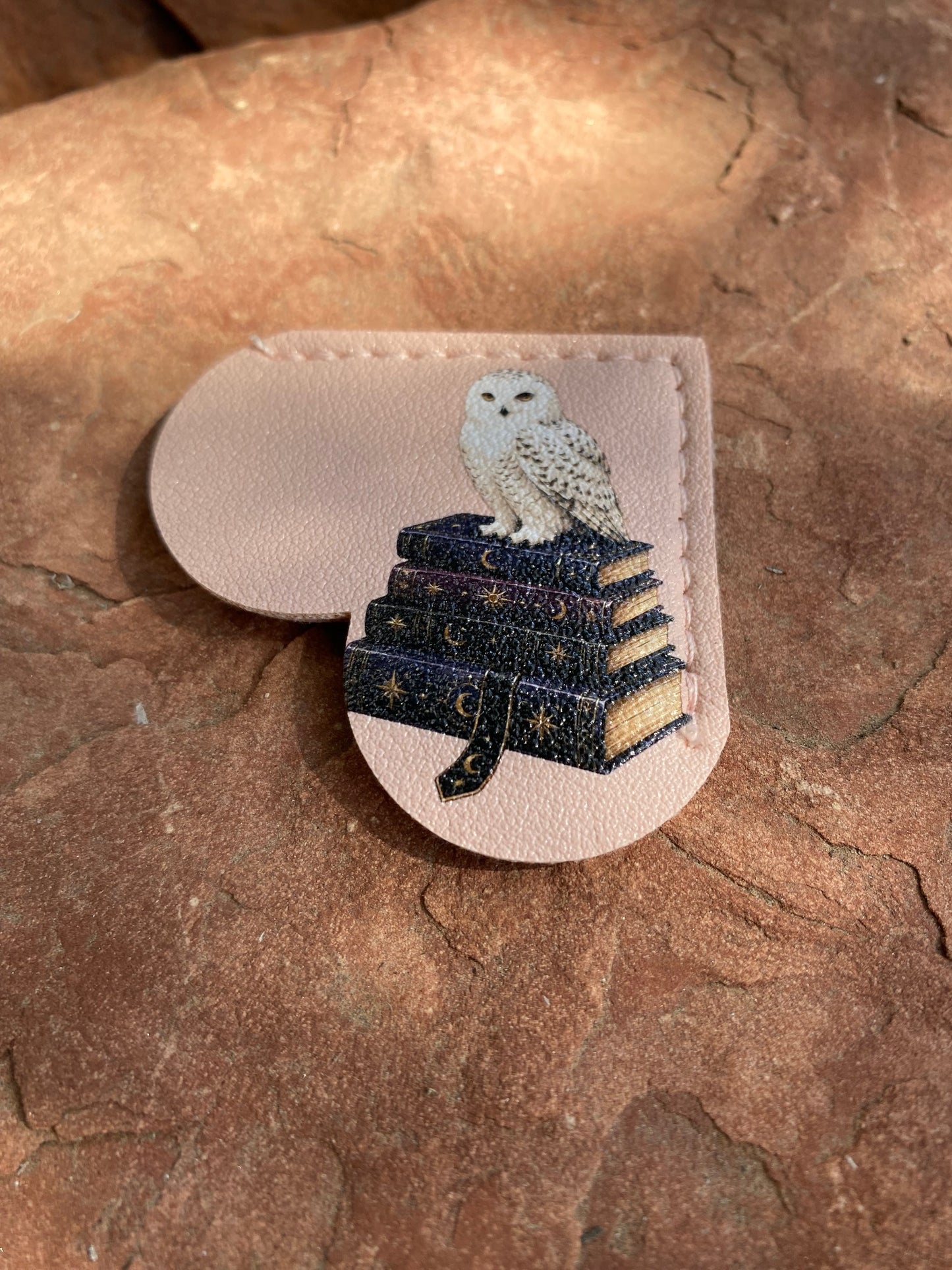 A small leather accessory with a stitched border and a printed illustration of a white owl sitting on stacked dark blue books with gold stars, placed on a textured brown stone surface.