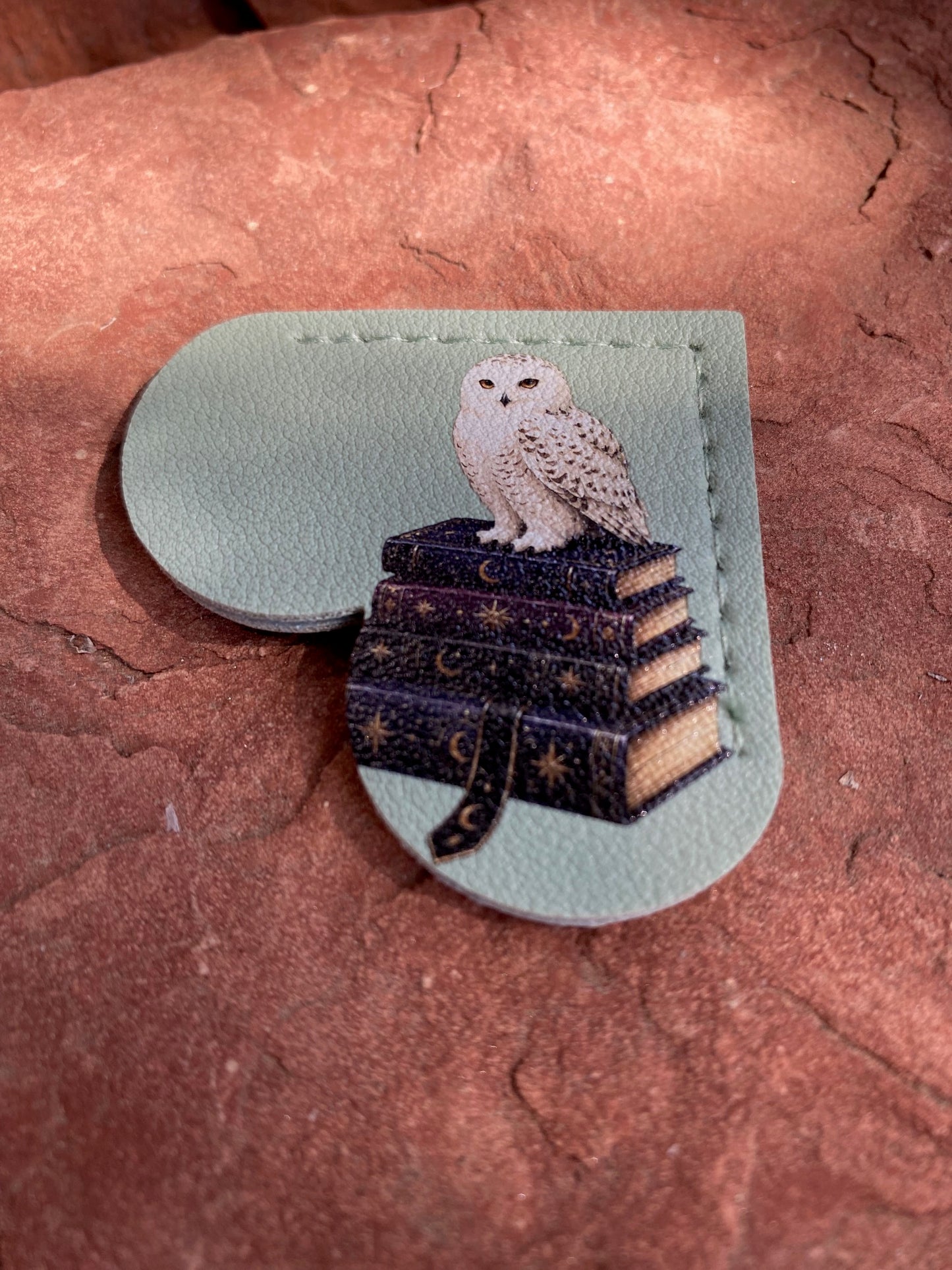 A small leather accessory with a stitched border and a printed illustration of a white owl perched on stacked dark books with gold stars, shown resting on a textured reddish-brown surface.