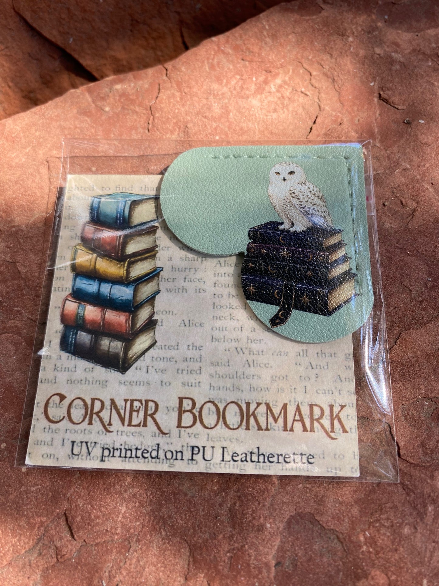 A packaged corner bookmark featuring a printed owl and books design on PU leatherette, shown in clear plastic wrapping on a reddish stone surface.