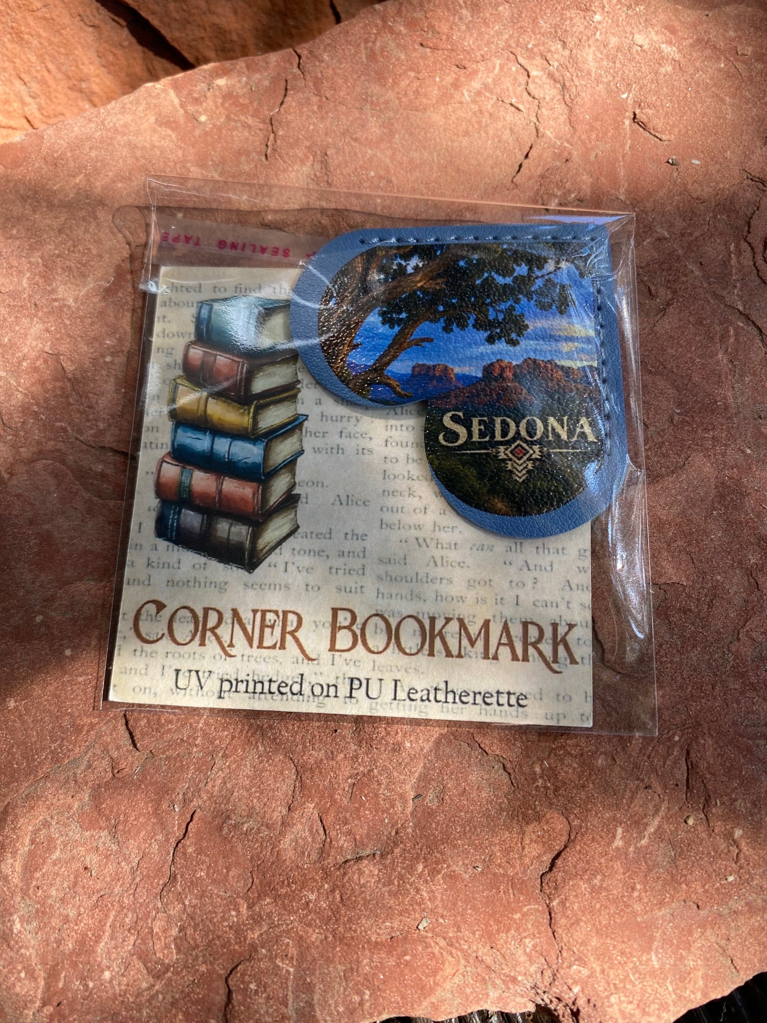 A packaged corner bookmark featuring a blue oval landscape patch with the word 'SEDONA' and an illustration of stacked books, shown on a red rock background.