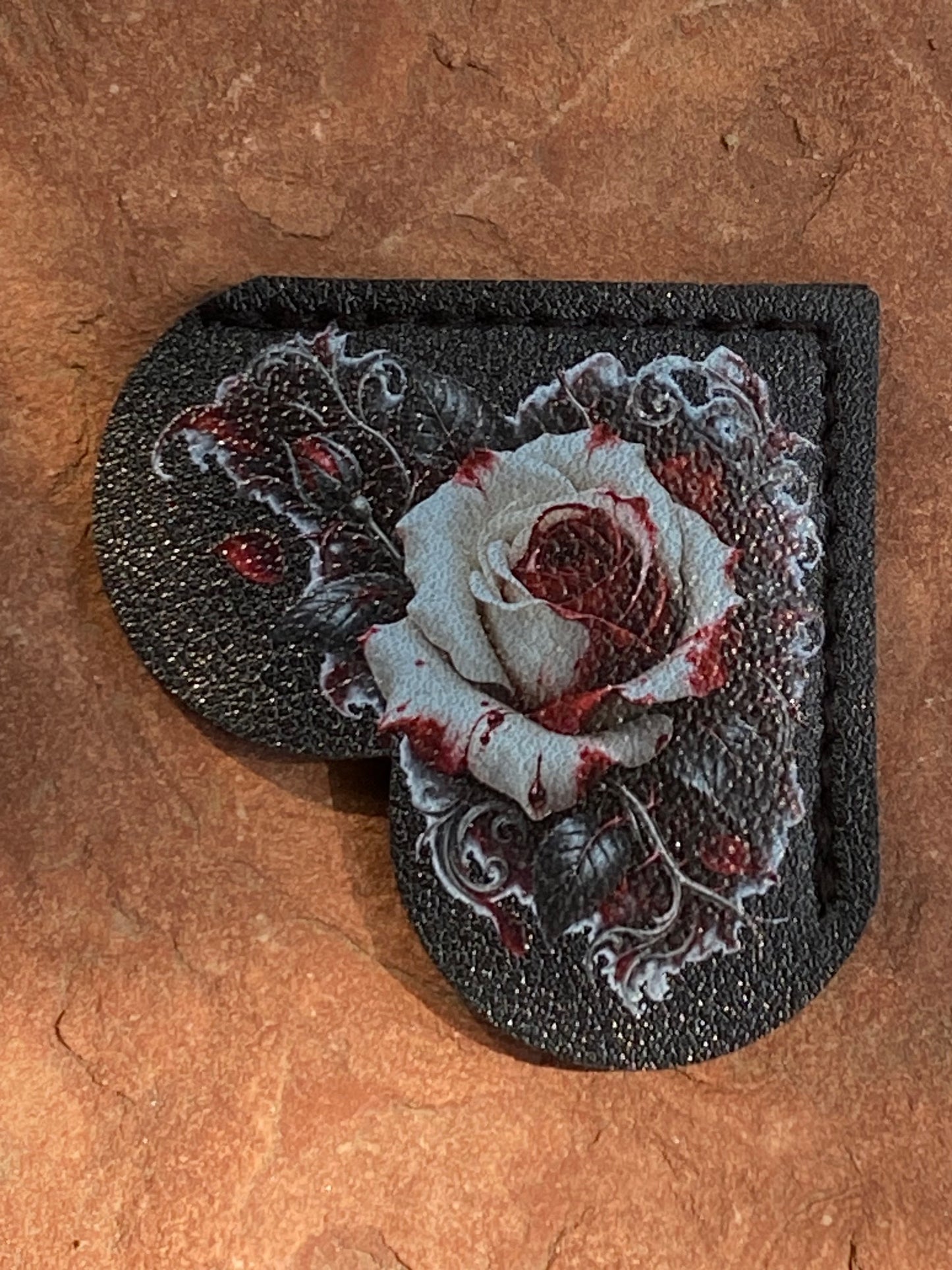 A heart-shaped black leather patch featuring a detailed embroidered rose design with white petals and red blood-like accents, displayed on a textured brown stone background.
