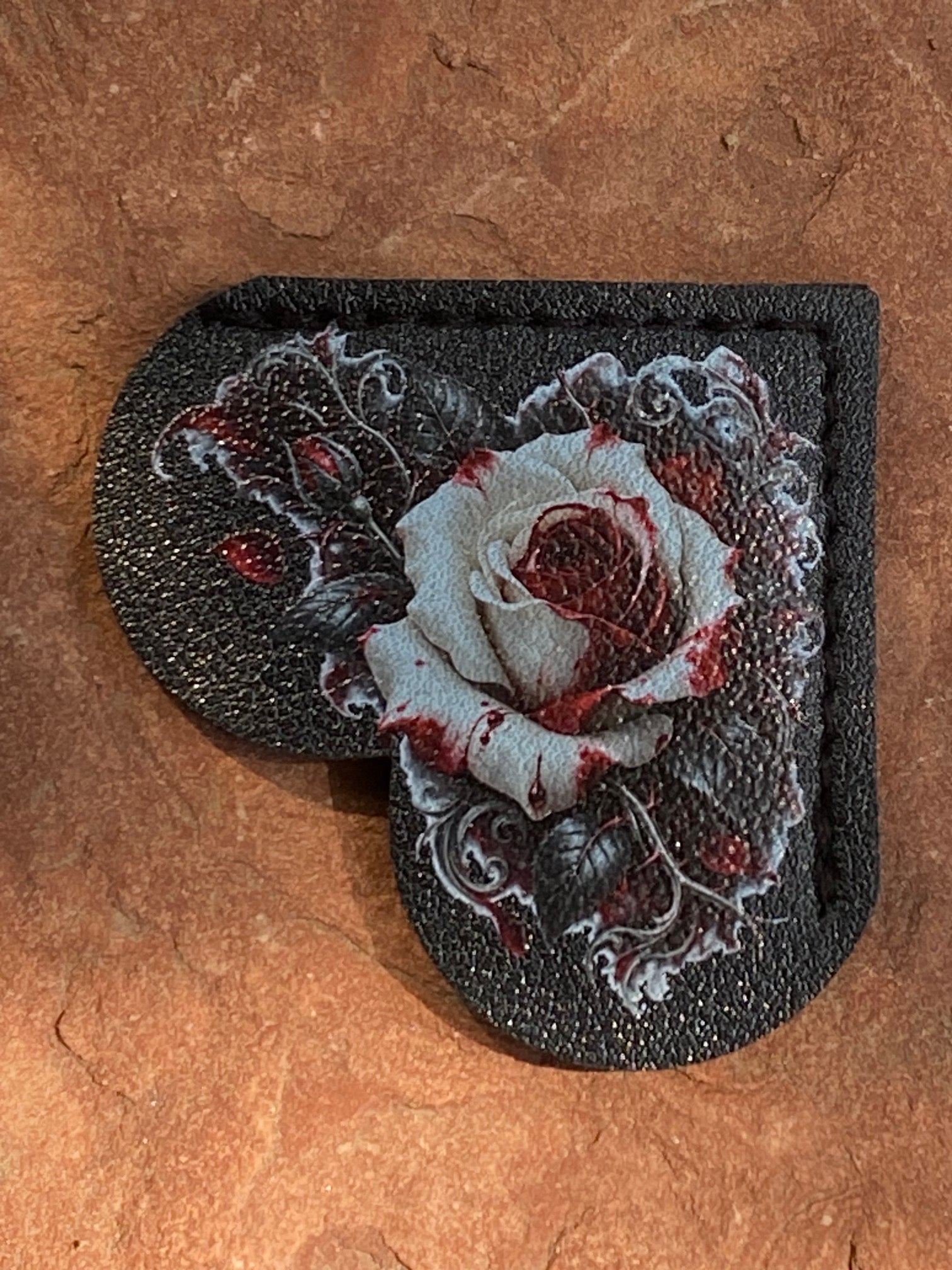 A heart-shaped black leather patch featuring a detailed embroidered rose design with white petals and red blood-like accents, displayed on a textured brown stone background.