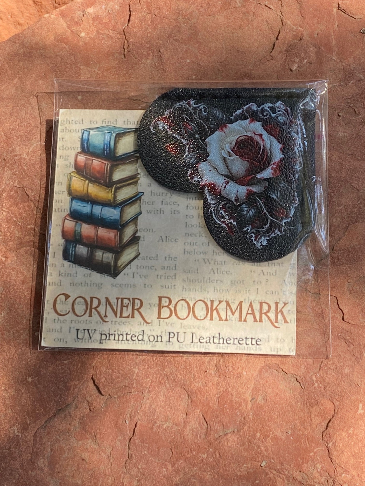 A packaged corner bookmark set on a textured terracotta surface. The set includes a rectangular card printed with vintage book stacks and the words 'CORNER BOOKMARK' and 'UV printed on PU Leatherette,' plus two oval leatherette corner pieces: one black with a white rose design and one black with a red rose design.