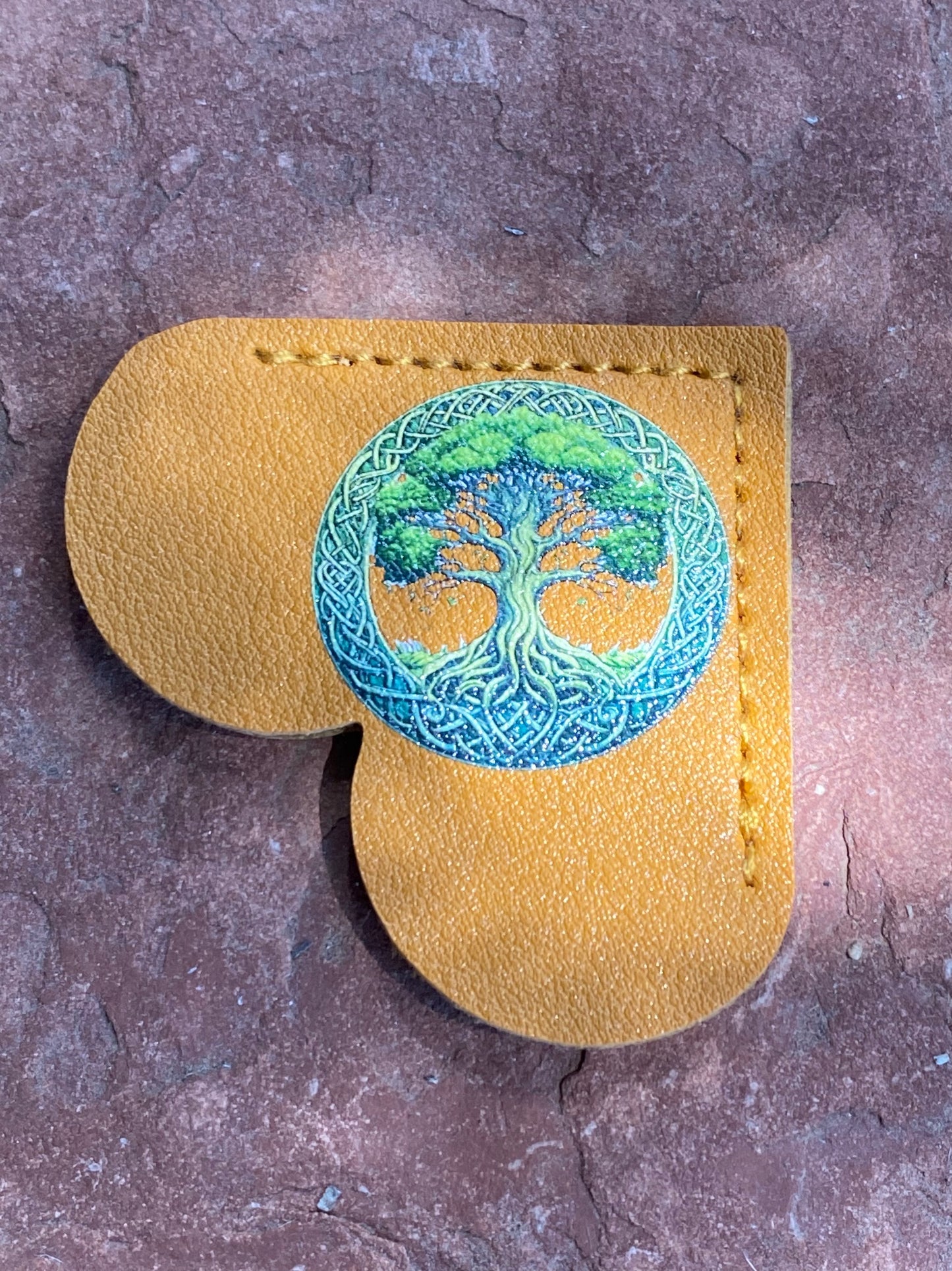 A mustard yellow leather pouch with a heart-shaped cutout, featuring a circular embroidered design of a green and blue tree of life on the front, displayed on a textured brown stone surface.