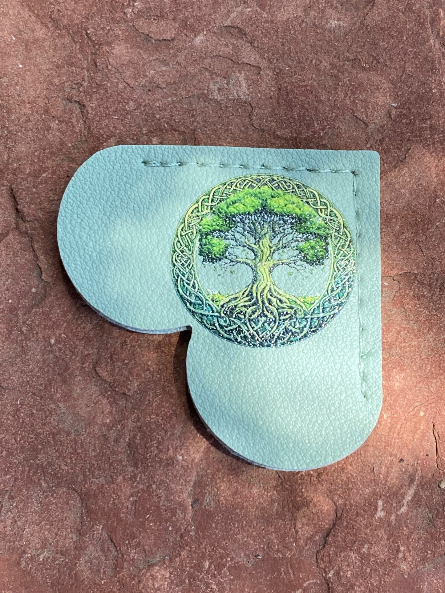 A light blue leather pouch with a heart-shaped cutout, featuring a green and yellow embroidered tree of life design on the front, set against a textured brown stone background.