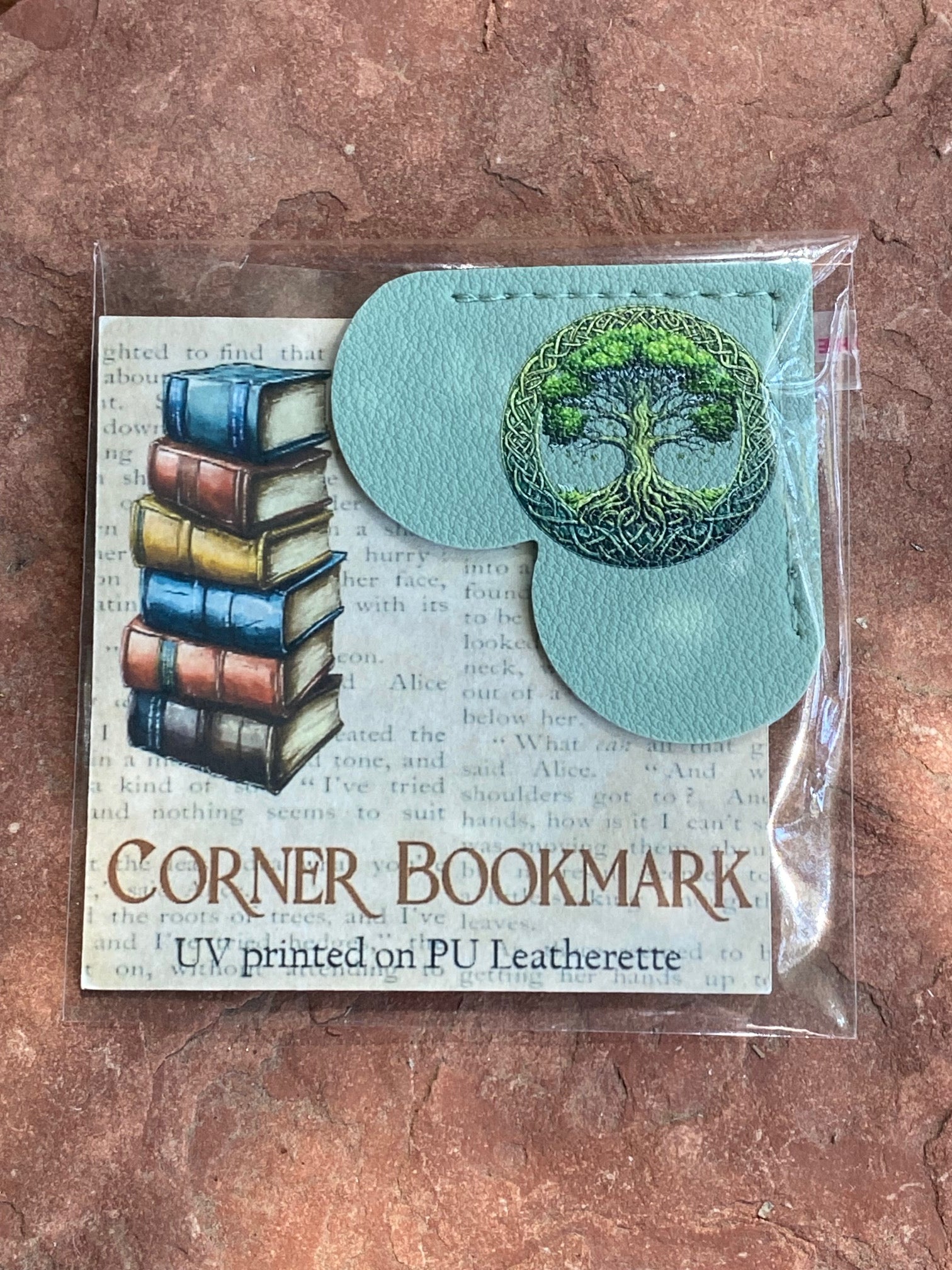 UV printed PU leatherette corner bookmarks in a clear plastic sleeve, displayed on a textured brown stone surface. The bookmarks feature a light teal color with one shaped like a stack of colorful books and the other shaped like a tree of life.