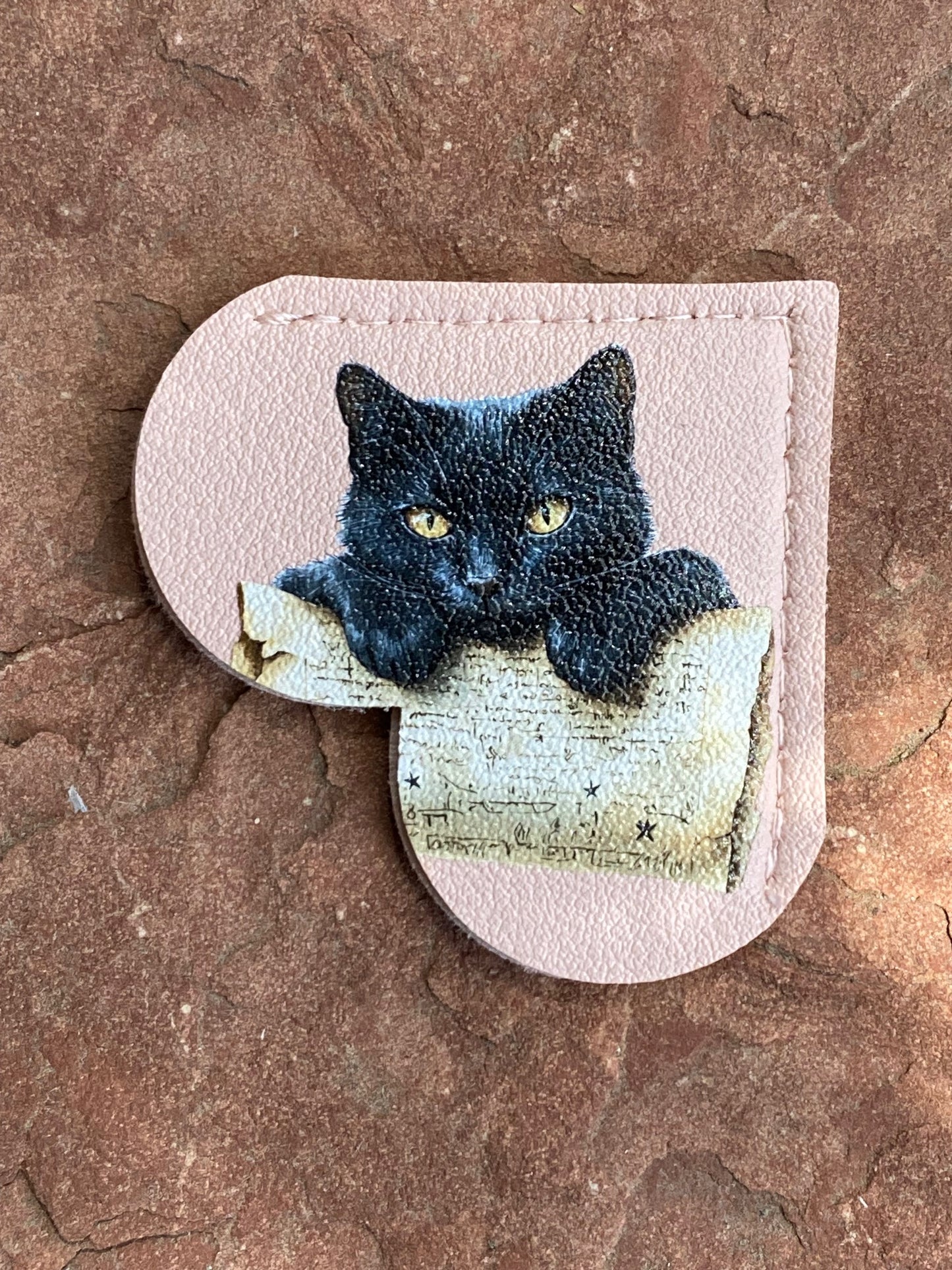 A light pink textured leather accessory, likely a card holder or wallet, featuring a printed illustration of a black cat with yellow eyes looking over an old book. The item is photographed flat on a textured brown stone surface.