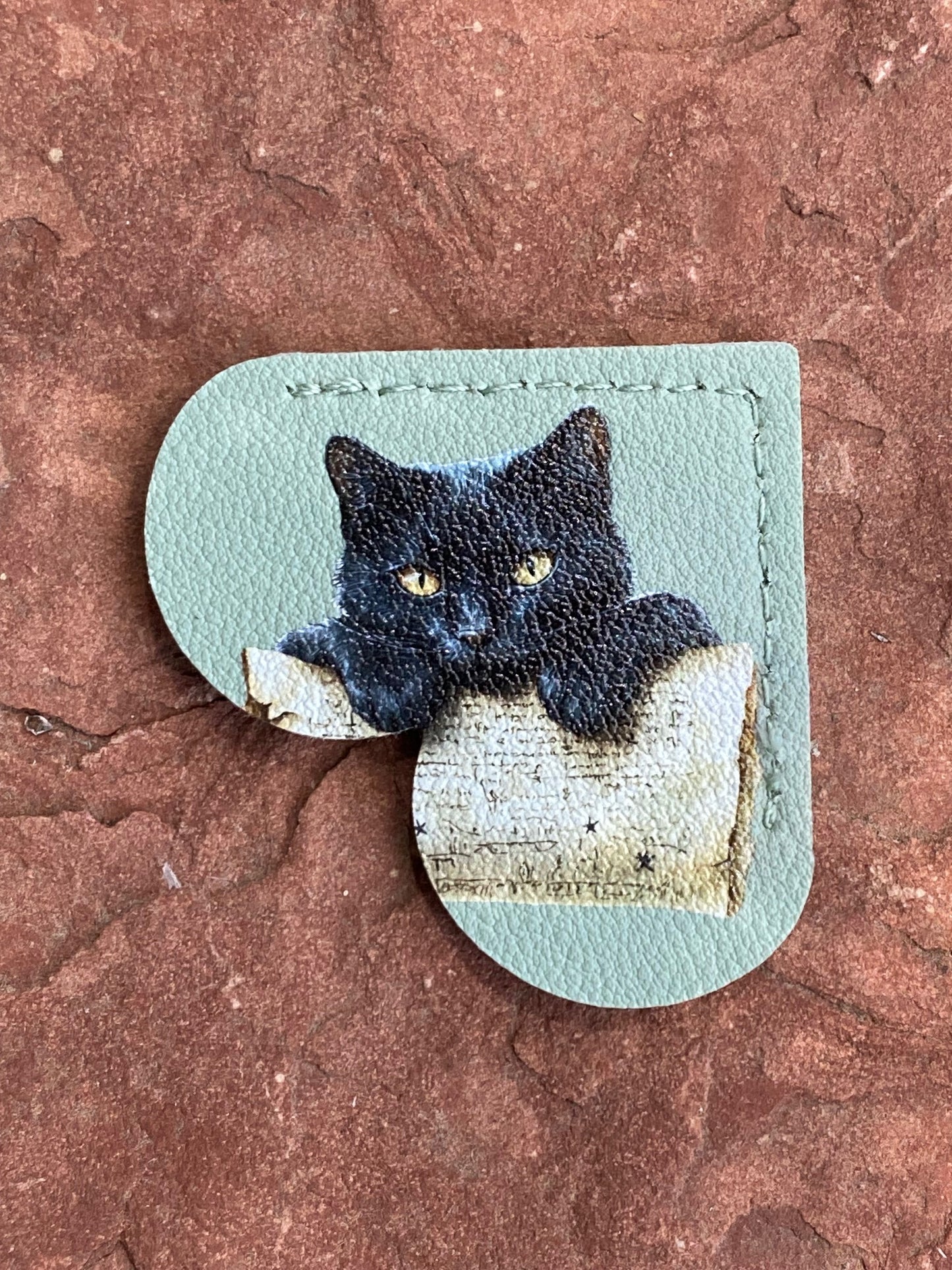 A handmade leather pouch featuring a printed illustration of a black cat peeking over a book on the front, set against a textured reddish-brown stone background.