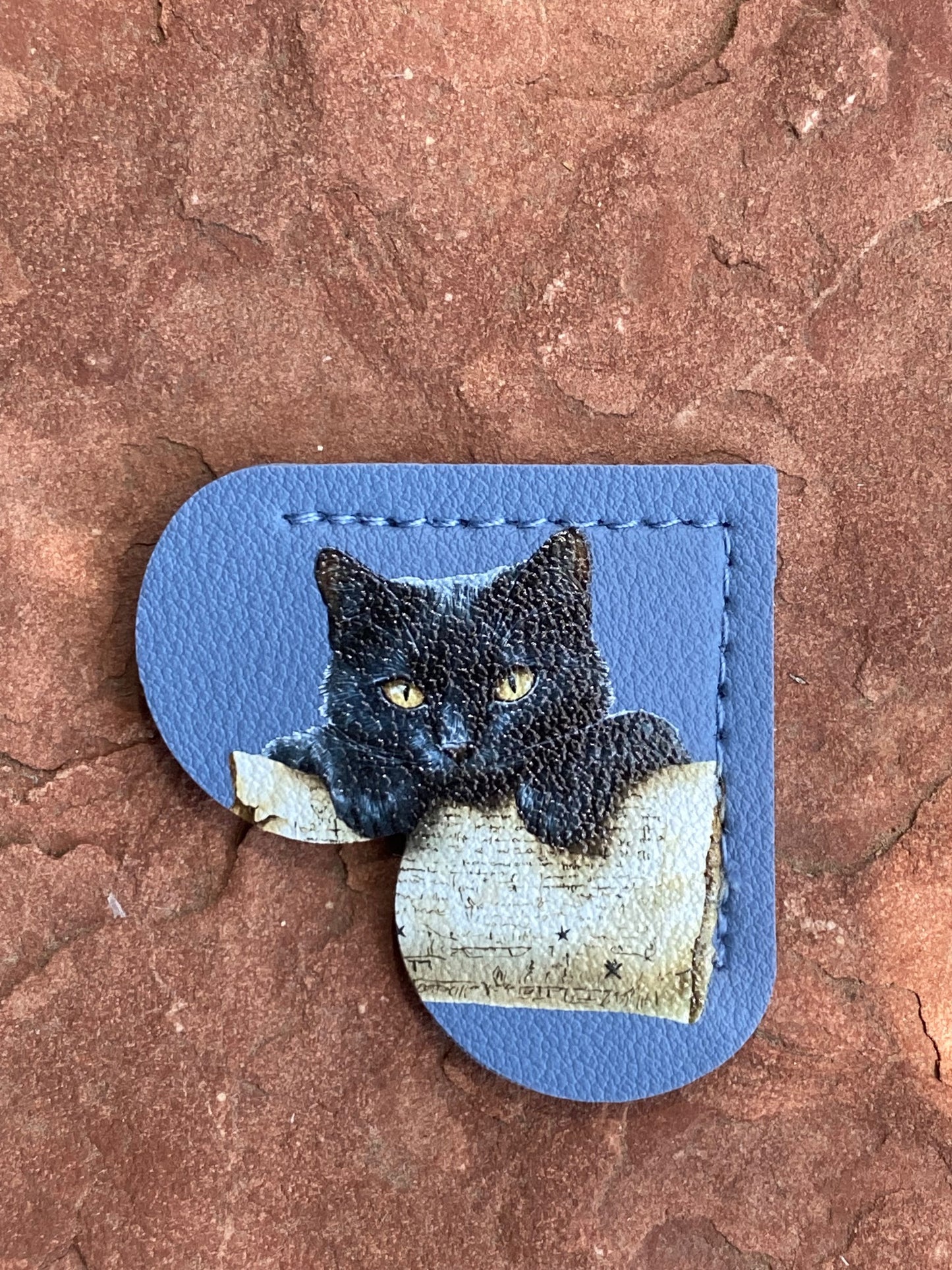 A blue leather patch featuring a printed black cat with yellow eyes looking over a book, displayed on a textured reddish-brown stone surface.