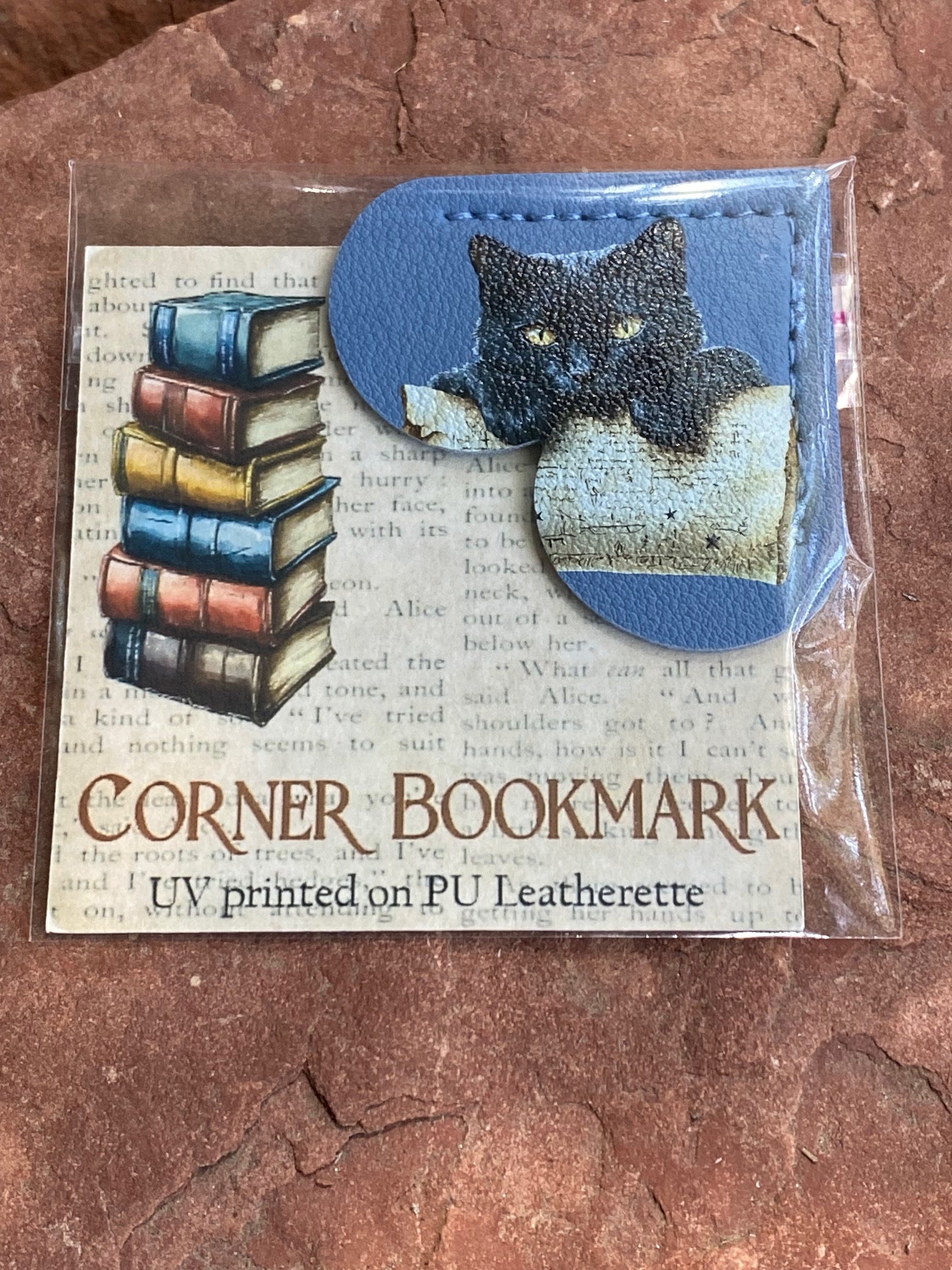A packaged corner bookmark featuring a blue leatherette tab with a black cat illustration and a stack of colorful books printed on a book-page background, placed on a textured reddish-brown surface.