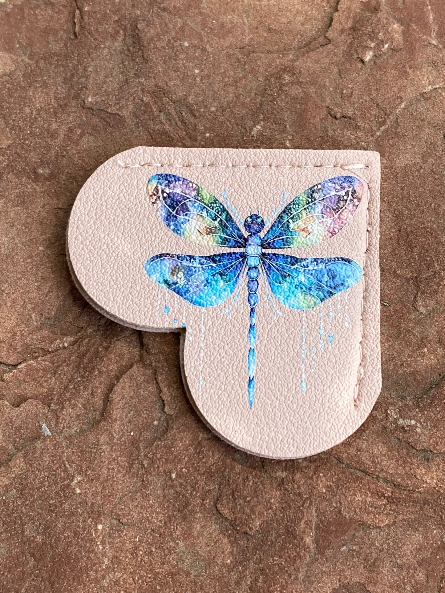 A light beige faux leather patch featuring a vibrant blue and multicolor watercolor dragonfly design, displayed on a textured brown stone surface.