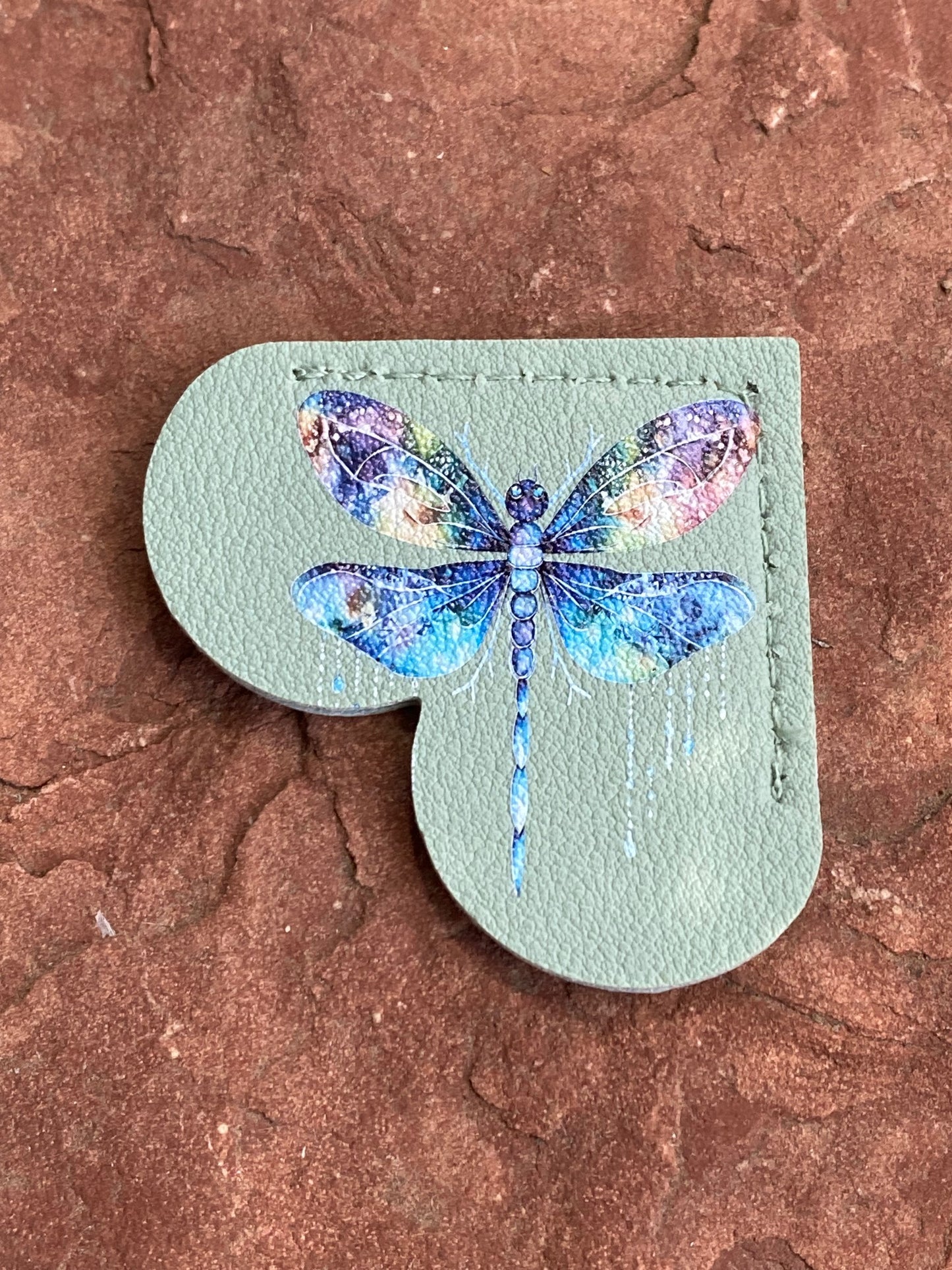 A heart-shaped sage green leather patch featuring a vibrant blue and purple watercolor dragonfly design, displayed on a textured reddish-brown stone surface.