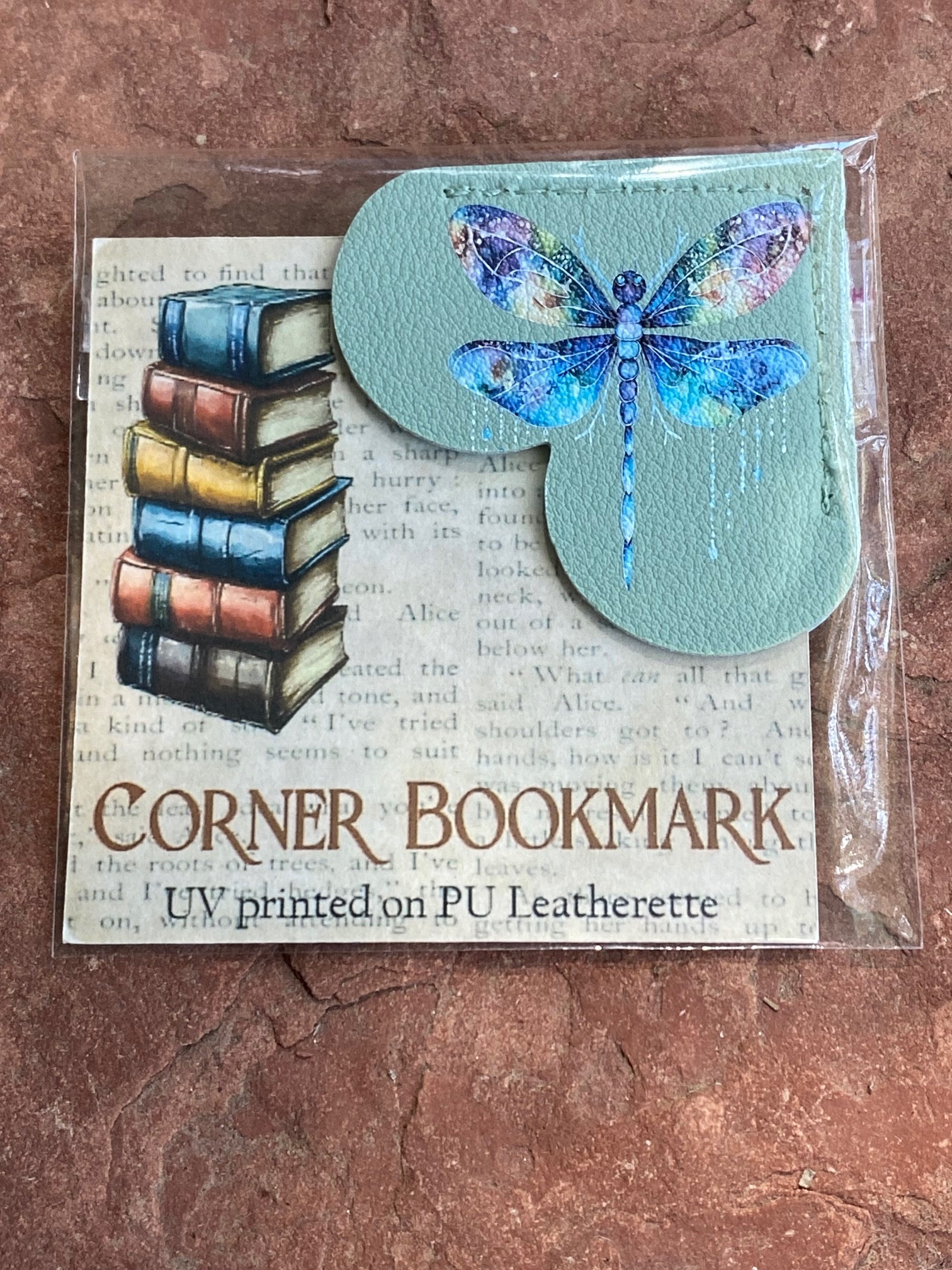 A packaged corner bookmark featuring a light blue-green PU leather shape with a colorful UV-printed dragonfly, shown on a book-print backing card with stacked books and the text 'CORNER BOOKMARK' and 'UV printed on PU Leatherette.'.