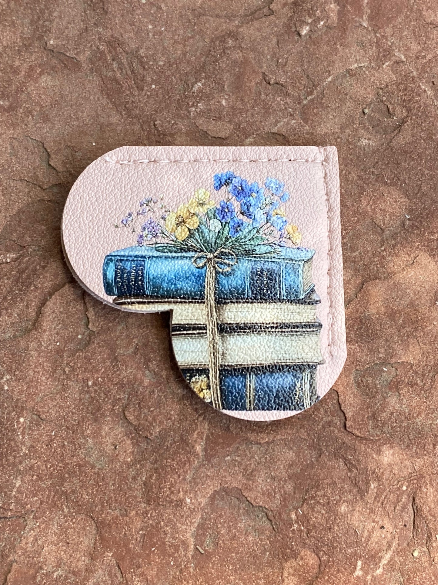 A heart-shaped leather accessory with a printed illustration of stacked blue books tied with a rope and topped with blue and yellow wildflowers, shown on a textured brown stone surface.
