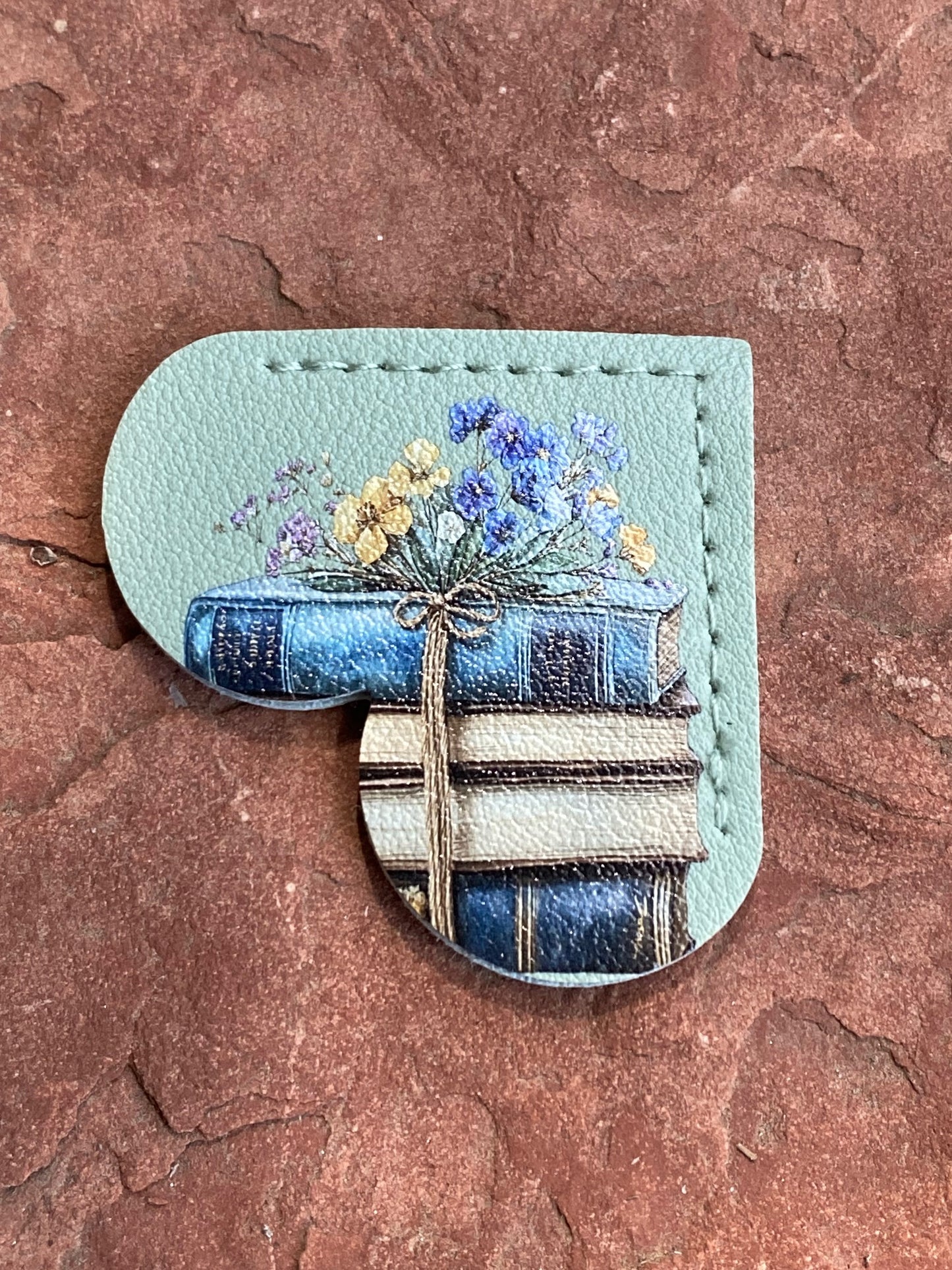 A heart-shaped leather patch featuring an illustration of stacked blue books tied with a ribbon and topped with small blue and yellow flowers, set against a textured reddish-brown stone background.