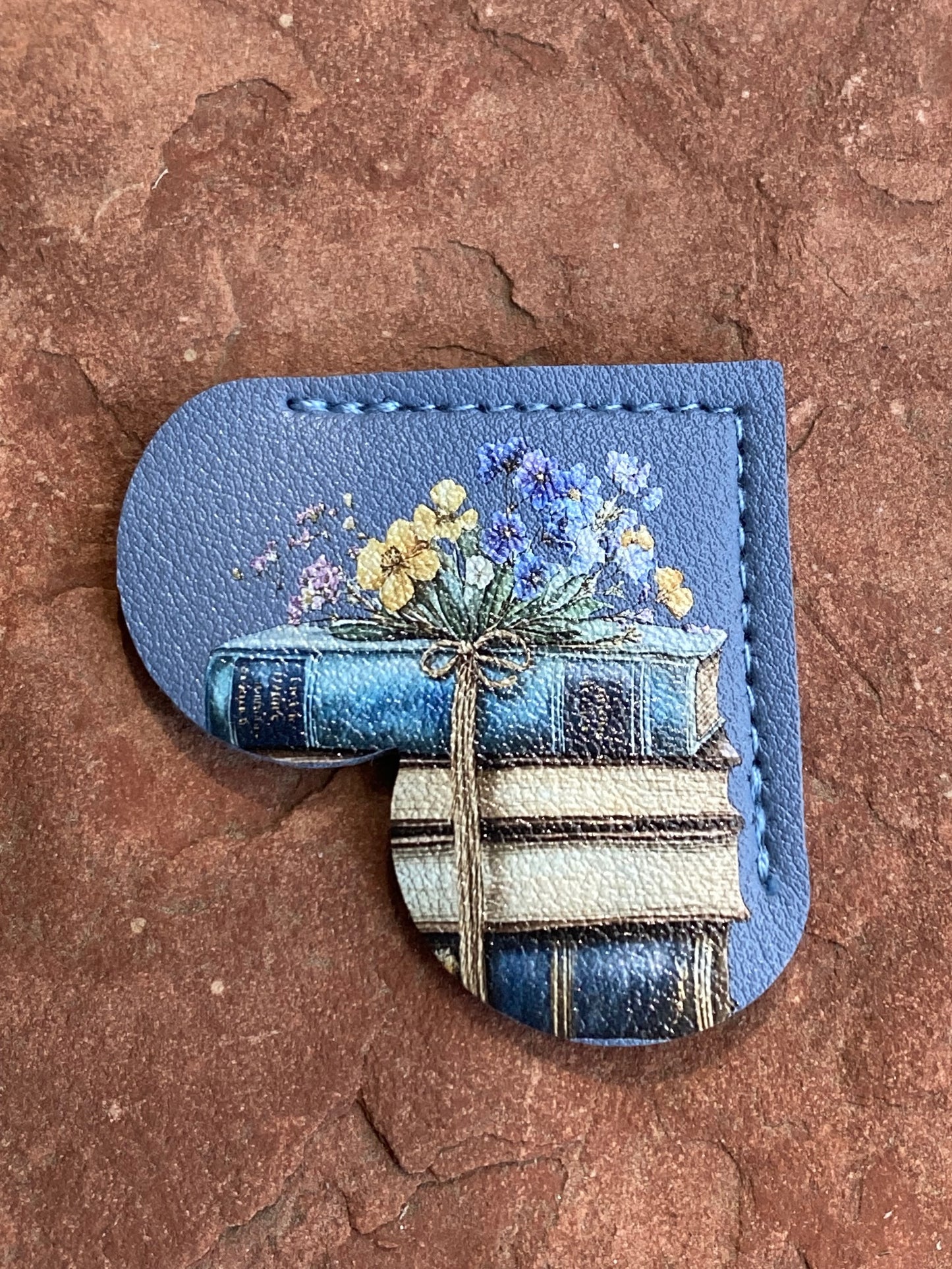 A blue leather-shaped patch or charm with a stitched border, featuring an illustration of stacked books with wildflowers on top, placed on a textured reddish-brown stone surface.