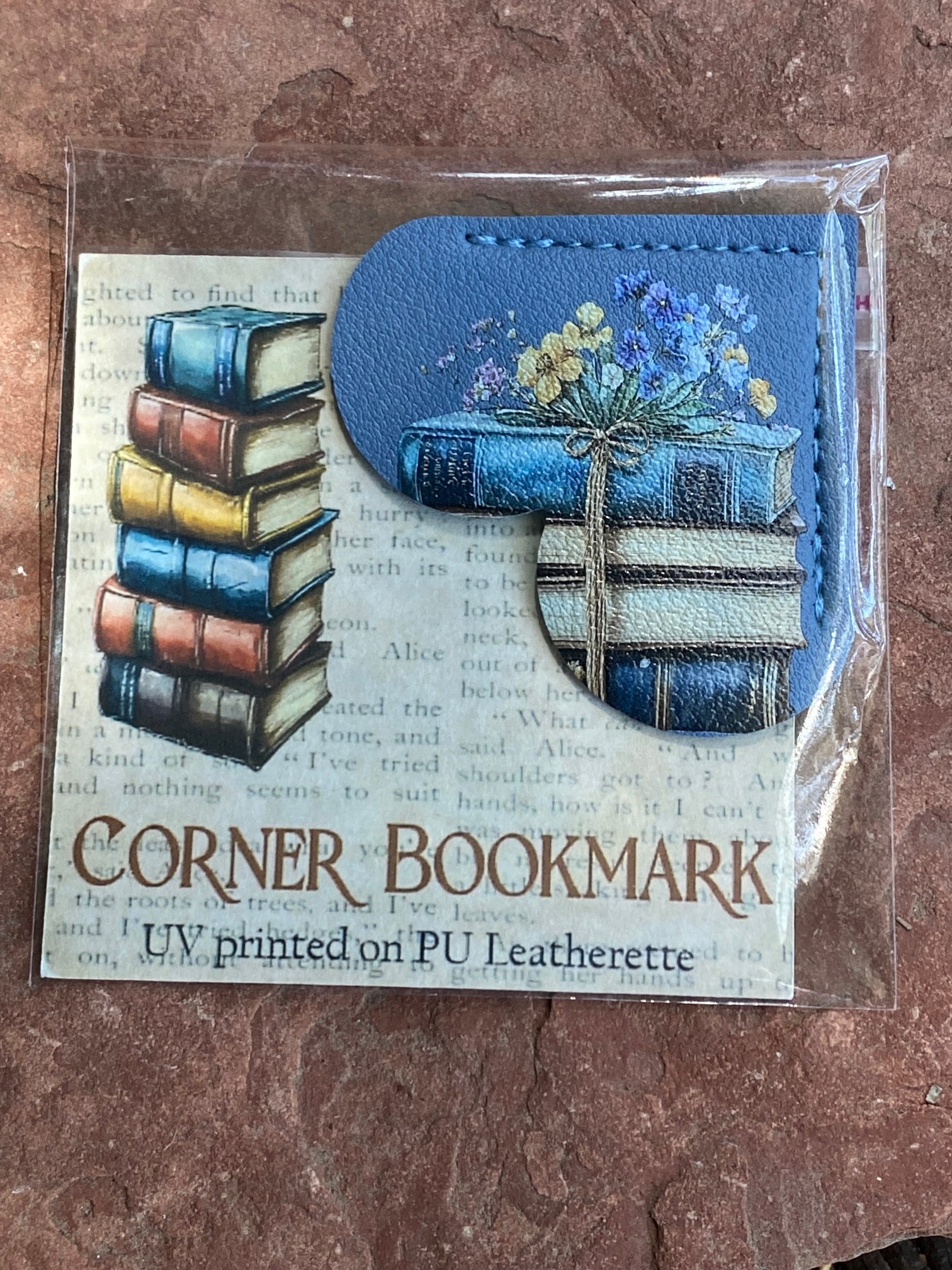 A packaged corner bookmark on a textured brown surface. The bookmark is blue faux leather with a printed design of stacked books and blue flowers, and the backing card reads 'CORNER BOOKMARK' and 'UV printed on PU Leatherette.