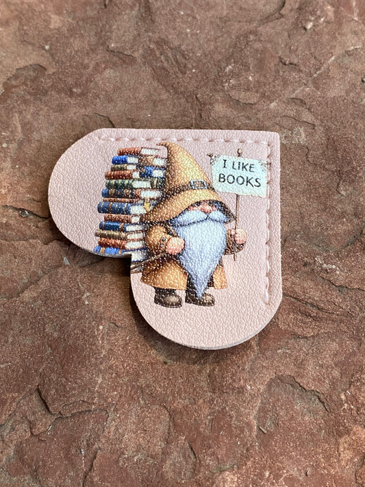 A heart-shaped beige leather patch featuring an illustration of a wizard gnomes holding a stack of books and a sign that reads 'I LIKE BOOKS', displayed on a textured brown stone surface.