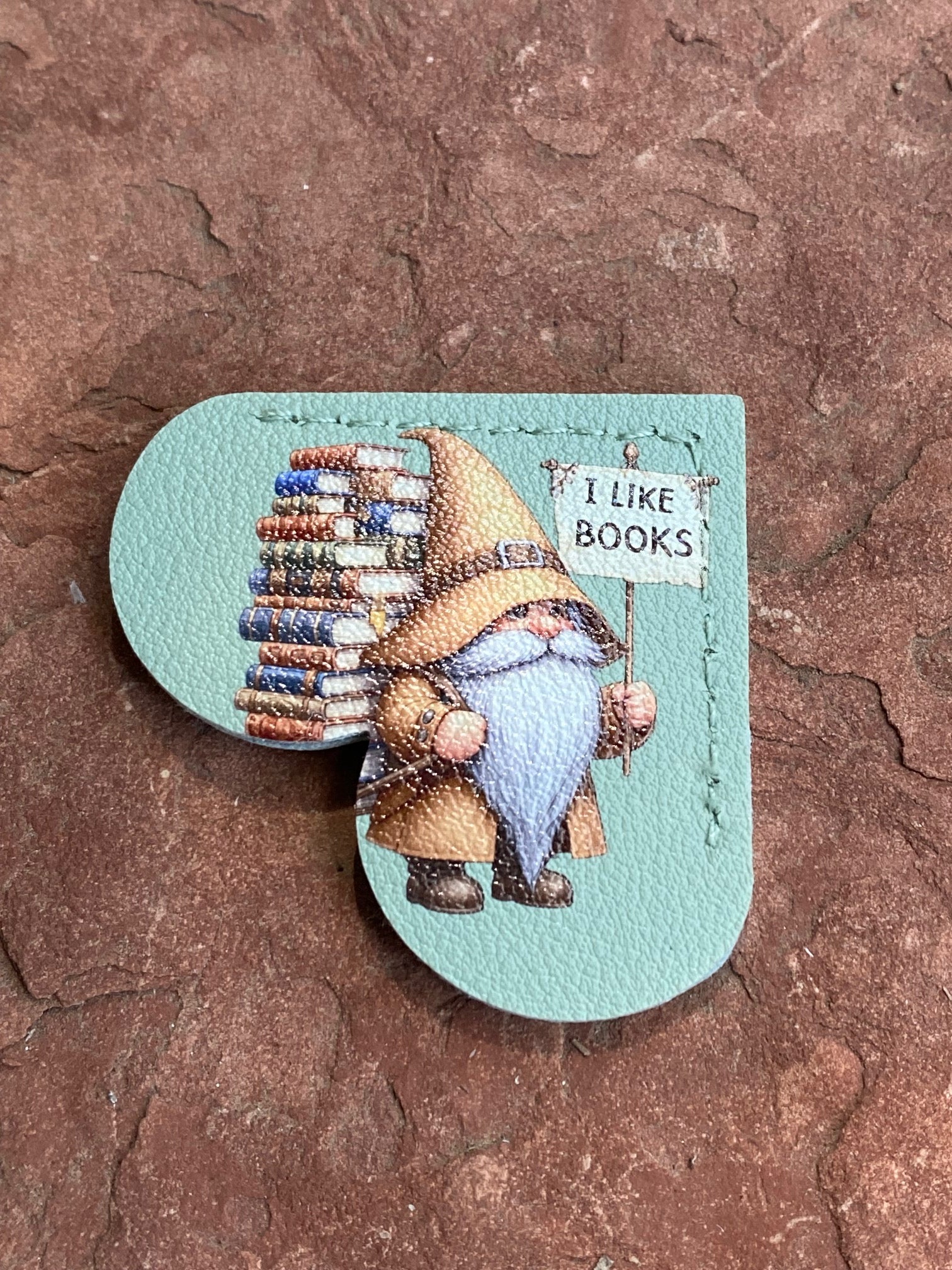 A heart-shaped leather bookmark with a stitched illustration of a book-loving gnome holding a sign that says 'I LIKE BOOKS', placed on a textured brown stone surface.