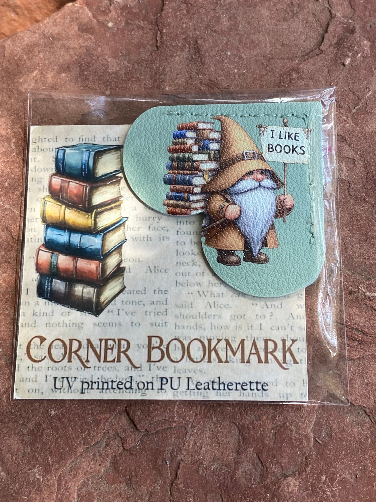 UV printed PU leatherette corner bookmark featuring a whimsical book-themed gnome illustration, shown in its clear plastic packaging on a textured brown stone surface.